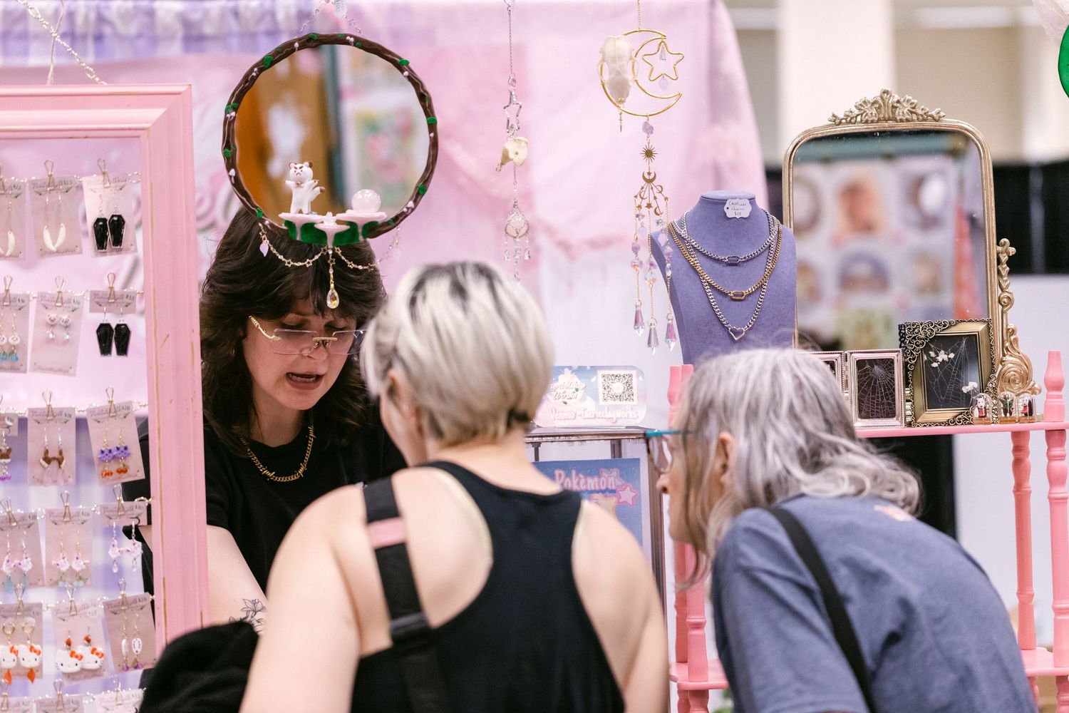 Woman selling jewelry at a pink booth display to customers at an indoor market.