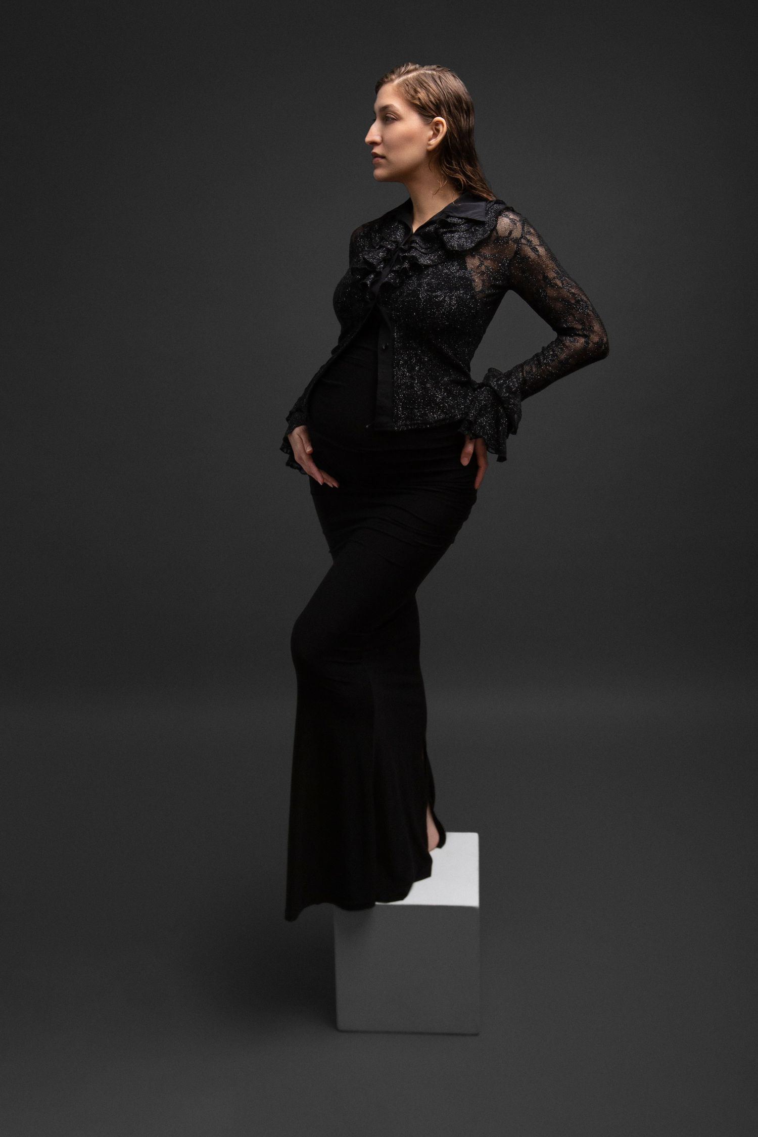 Woman in elegant black lace blouse and flared skirt posing on white cube against dark gray background.