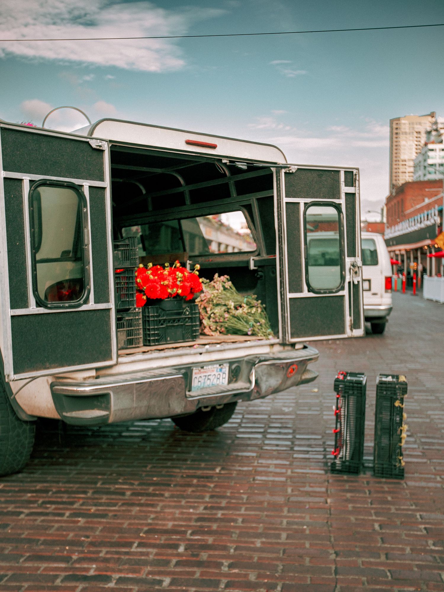 Delivery truck with open rear doors loaded with red flowers and greenery on a brick street market.
