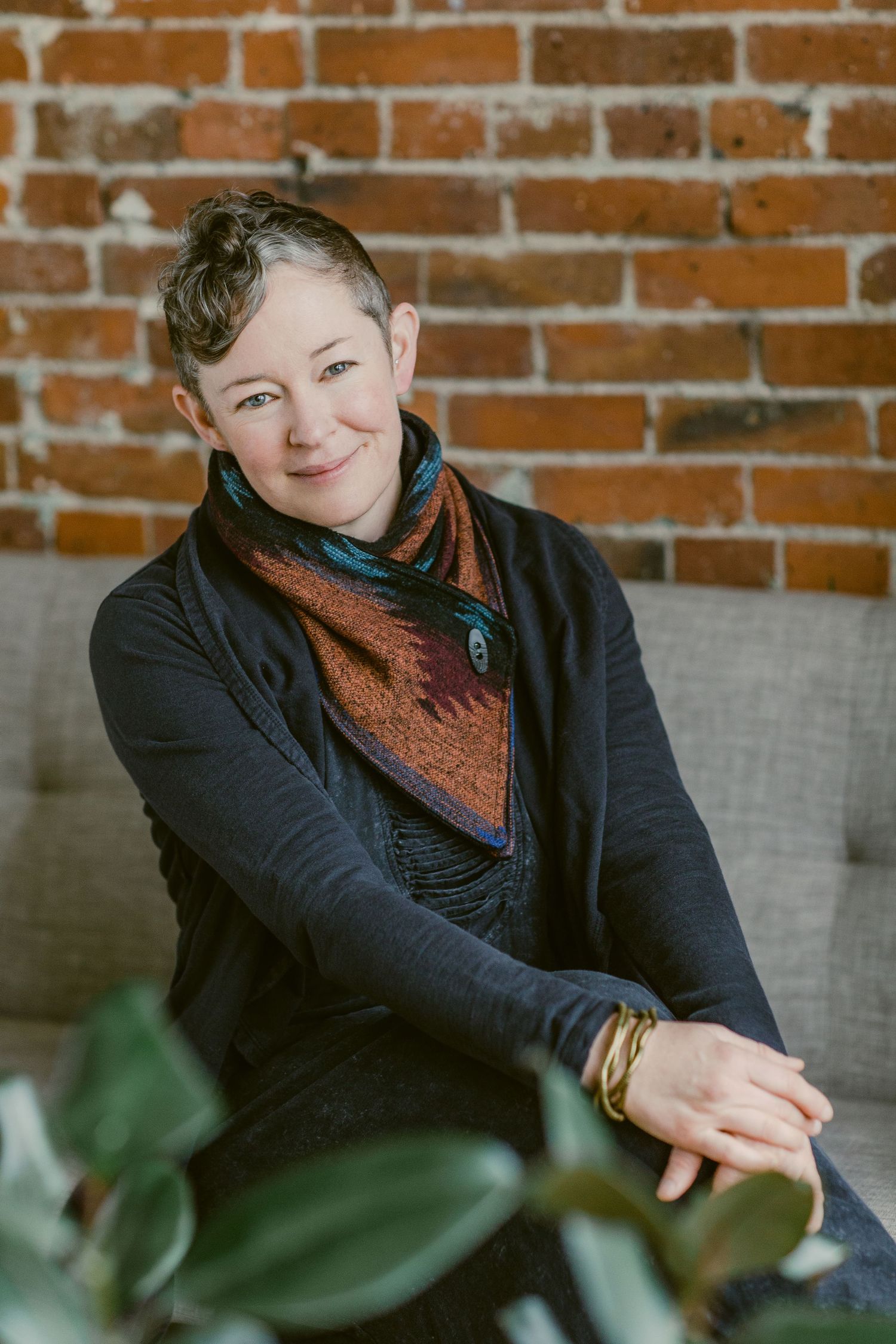 Smiling woman with short hair wearing a colorful scarf and dark cardigan, seated on a couch against a brick wall.
