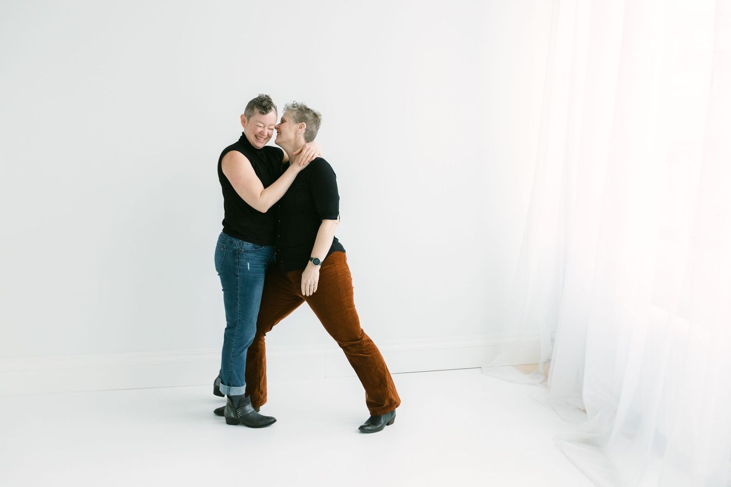 Two people dancing together in a bright white studio, wearing casual black tops and colorful pants.