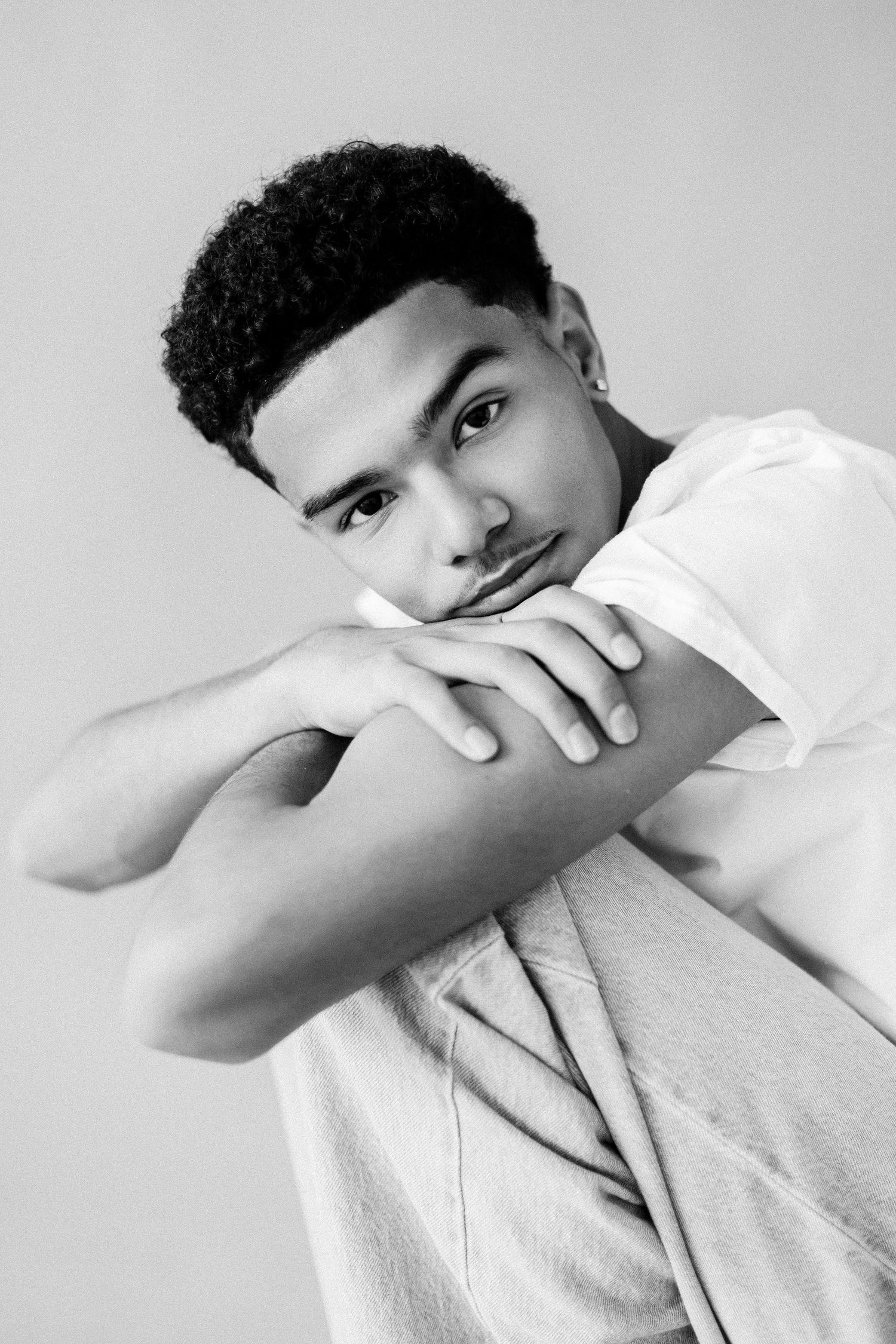 Black and white portrait of a young man in a white t-shirt posing with arms crossed, resting chin on hands.