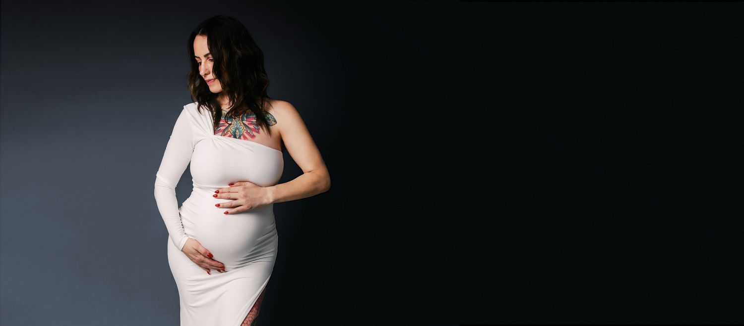Pregnant woman in white dress cradling baby bump against dark background in elegant maternity photo.