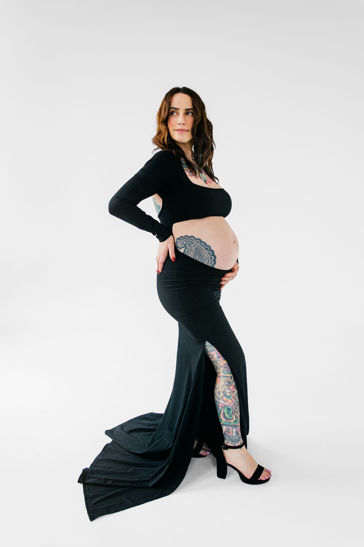 Pregnant woman in black crop top and maxi skirt with tattoos poses for maternity photo on white background.