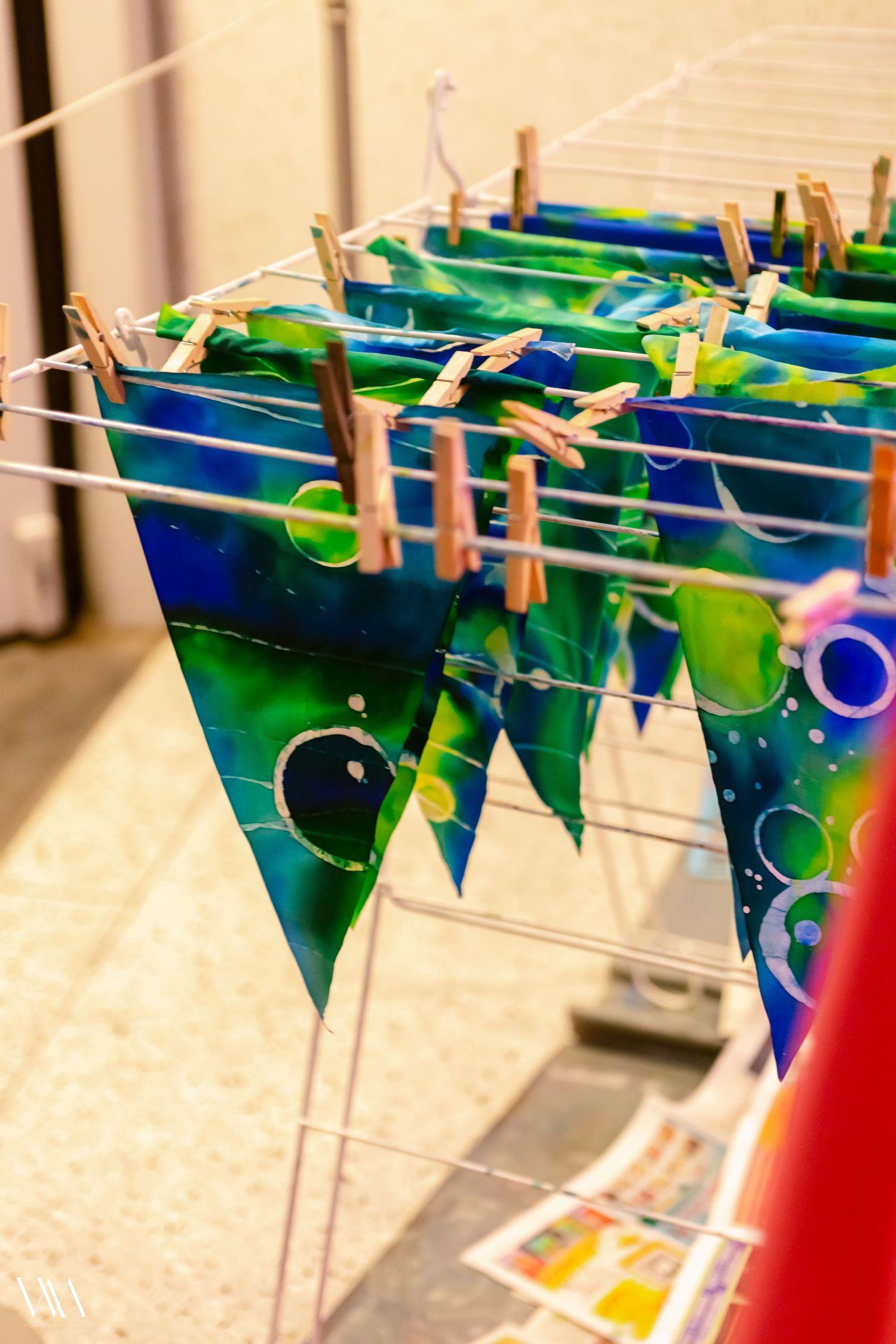 Colorful tie-dye triangular flags clipped with wooden clothespins on wire drying rack, featuring blue and green bubble patterns.
