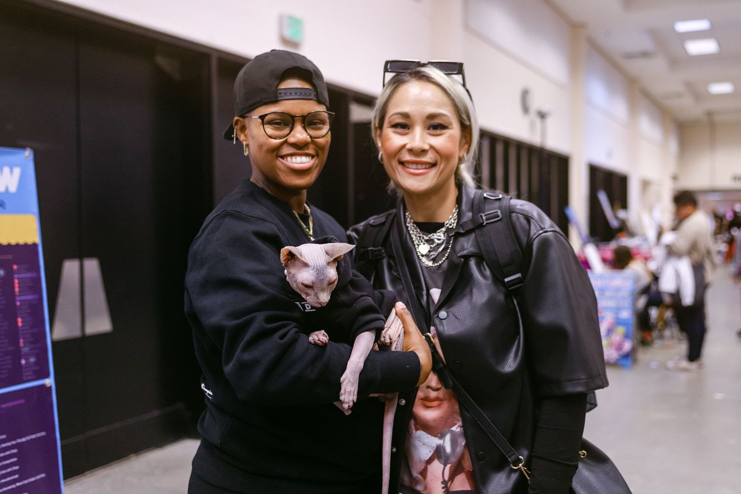 Two smiling women in black outfits pose together holding a hairless Sphynx cat at an indoor event.