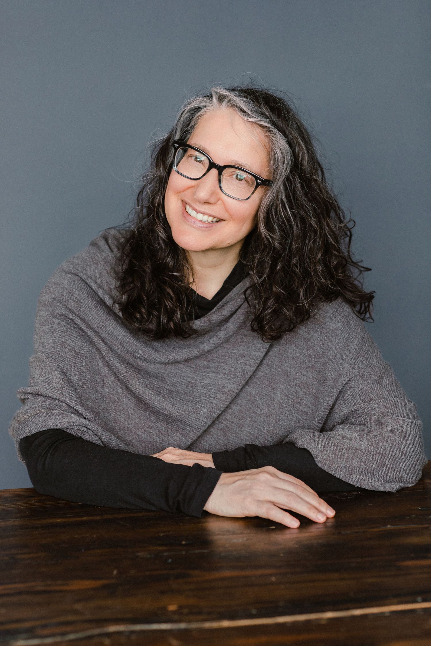 Smiling middle-aged woman with curly gray-black hair and black glasses, wearing gray wrap top, seated at wooden table.