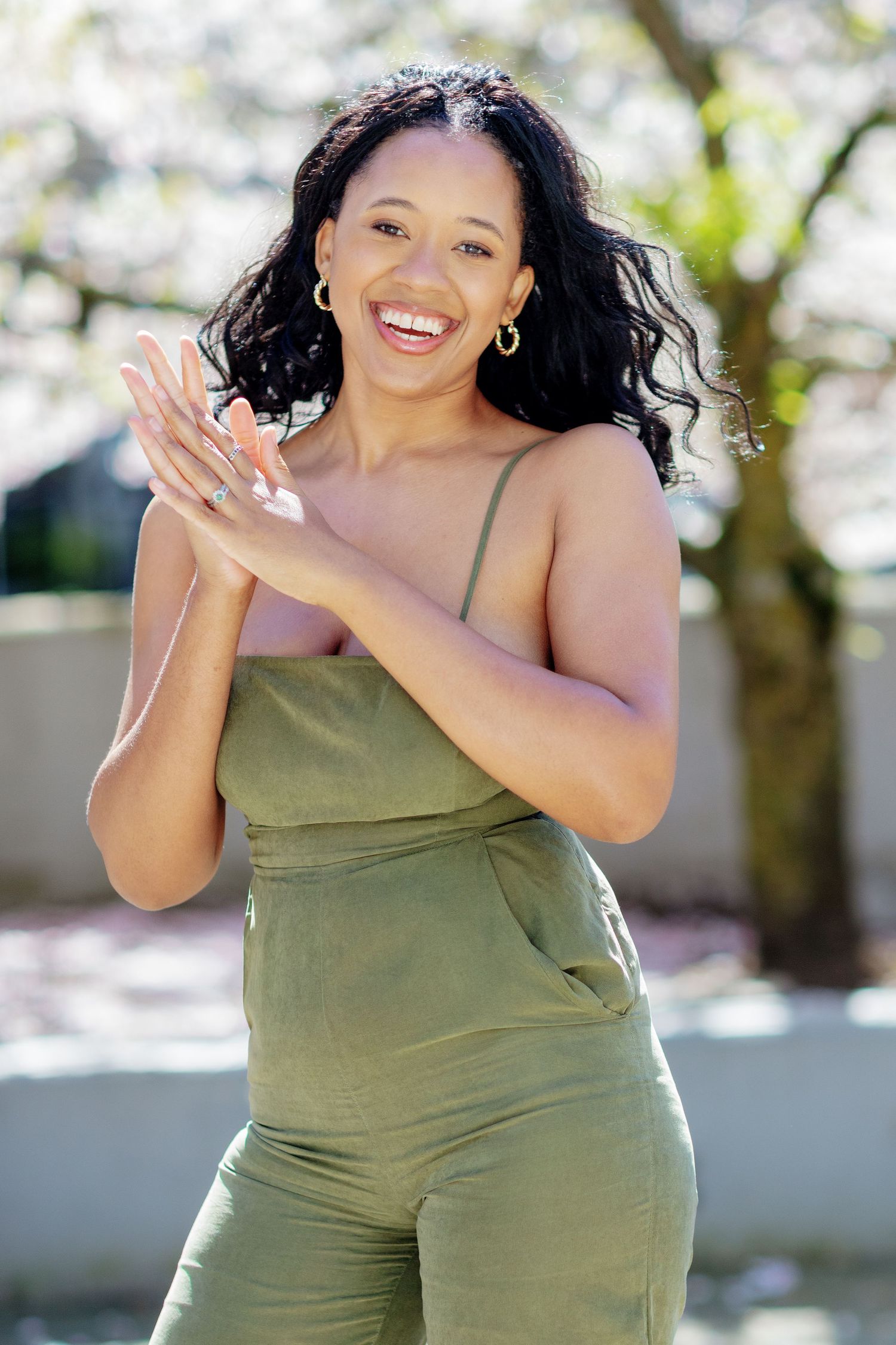 Joyful woman in olive green jumpsuit clapping hands, laughing outdoors near blooming cherry trees.