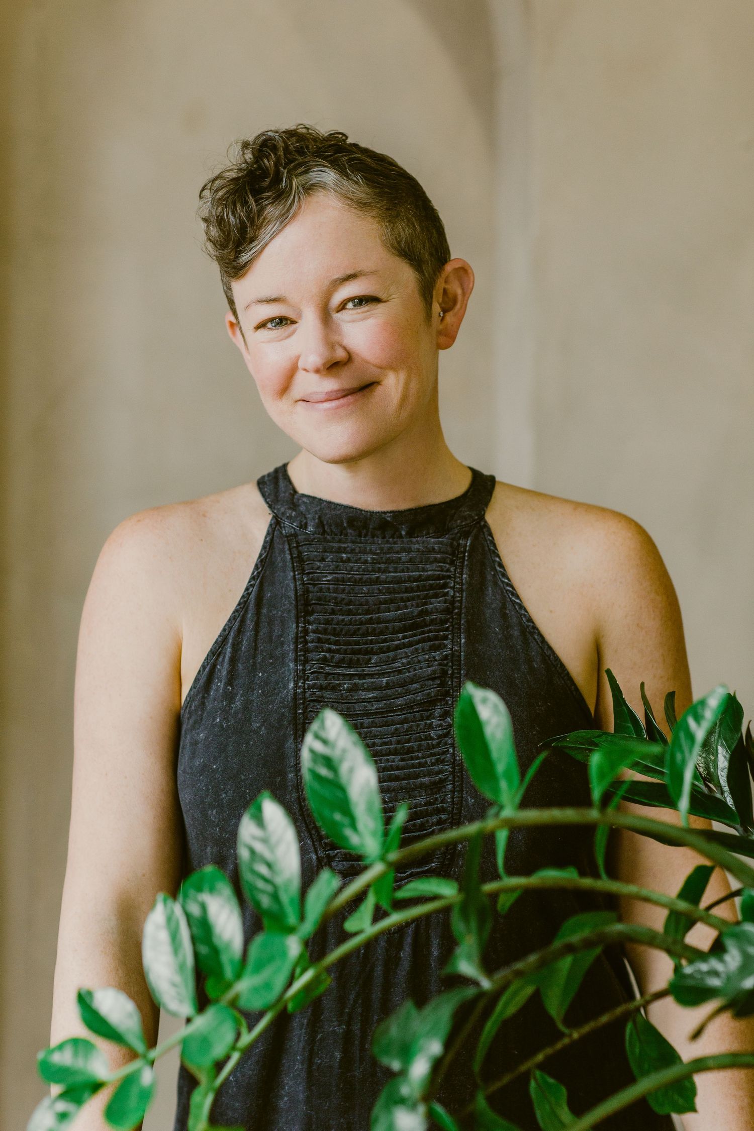 Smiling woman with short curly hair wearing a black halter top, standing near a green leafy plant indoors.