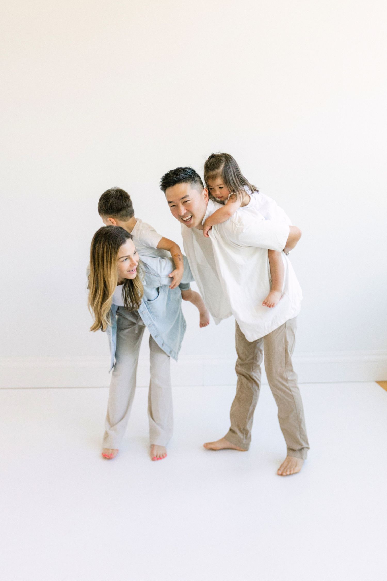 Happy Asian family of four playing piggyback rides together in a bright white studio setting.