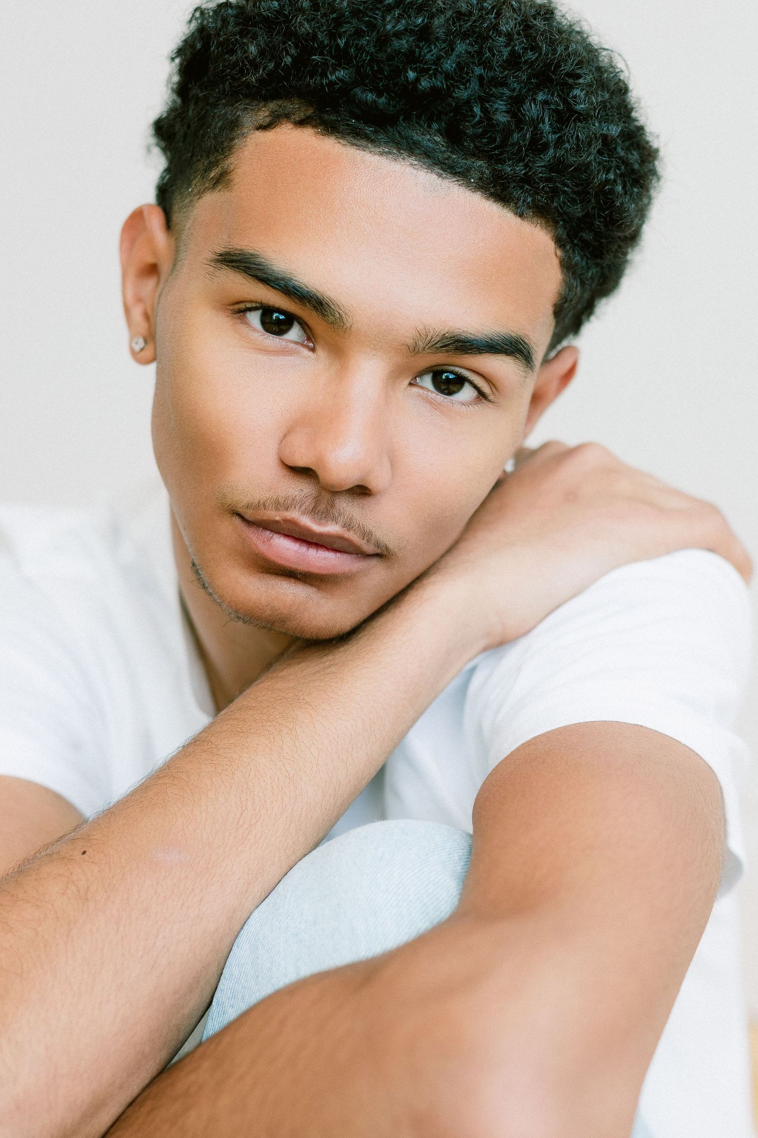 Young man with curly hair wearing a white t-shirt, resting his chin on his hand against a light background.