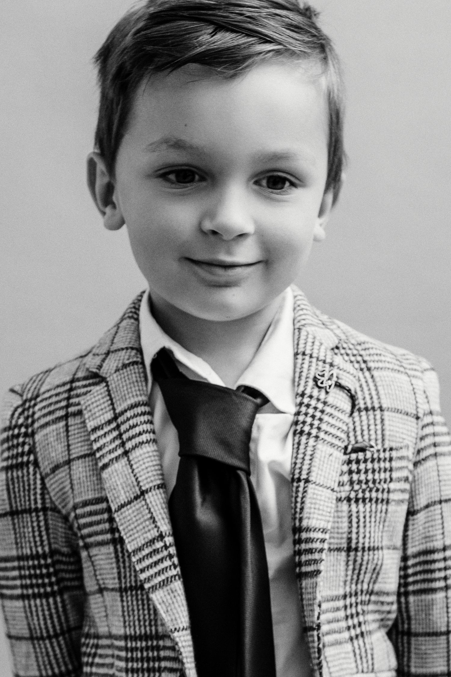 Black and white portrait of a young boy in a plaid blazer and dark tie, smiling confidently at the camera.