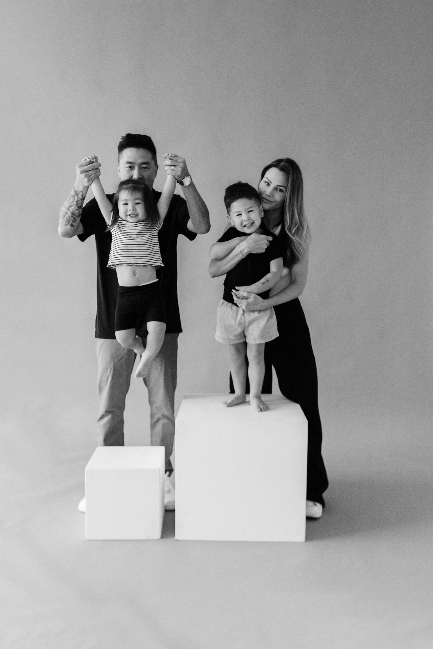 Black and white family portrait with parents holding two young children on white pedestals in a studio.