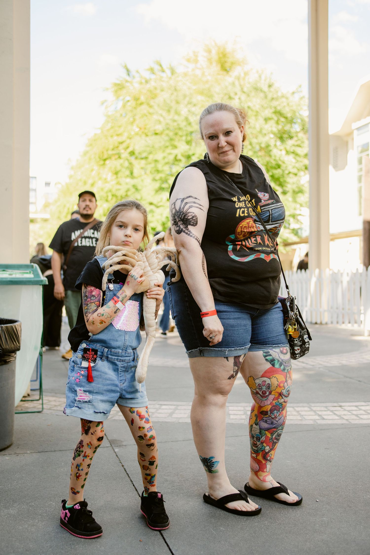 Heavily tattooed woman and young girl with fake tattoos posing together outdoors at a public event.