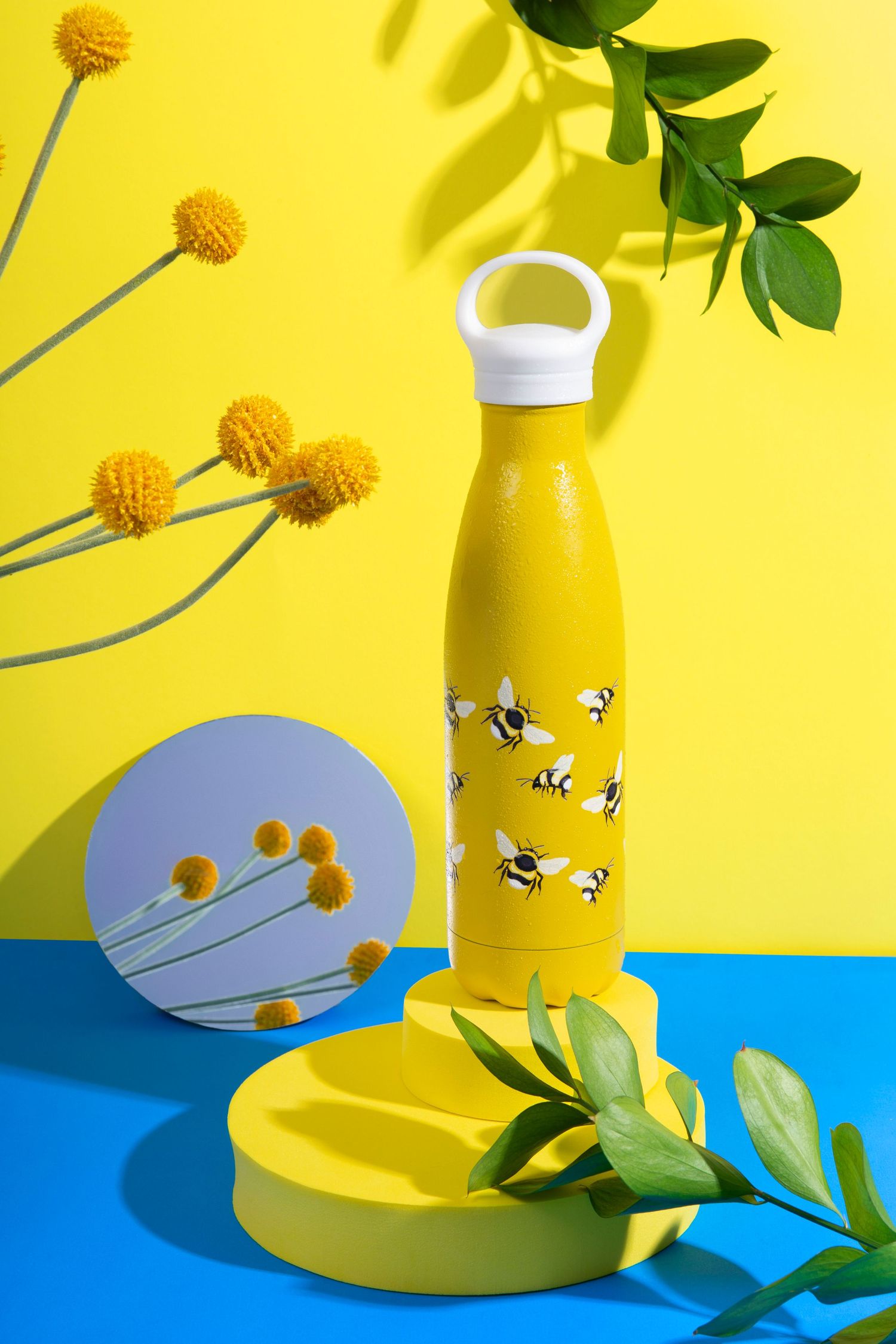 Yellow water bottle with bee print design on yellow podium, surrounded by yellow flowers and green leaves.