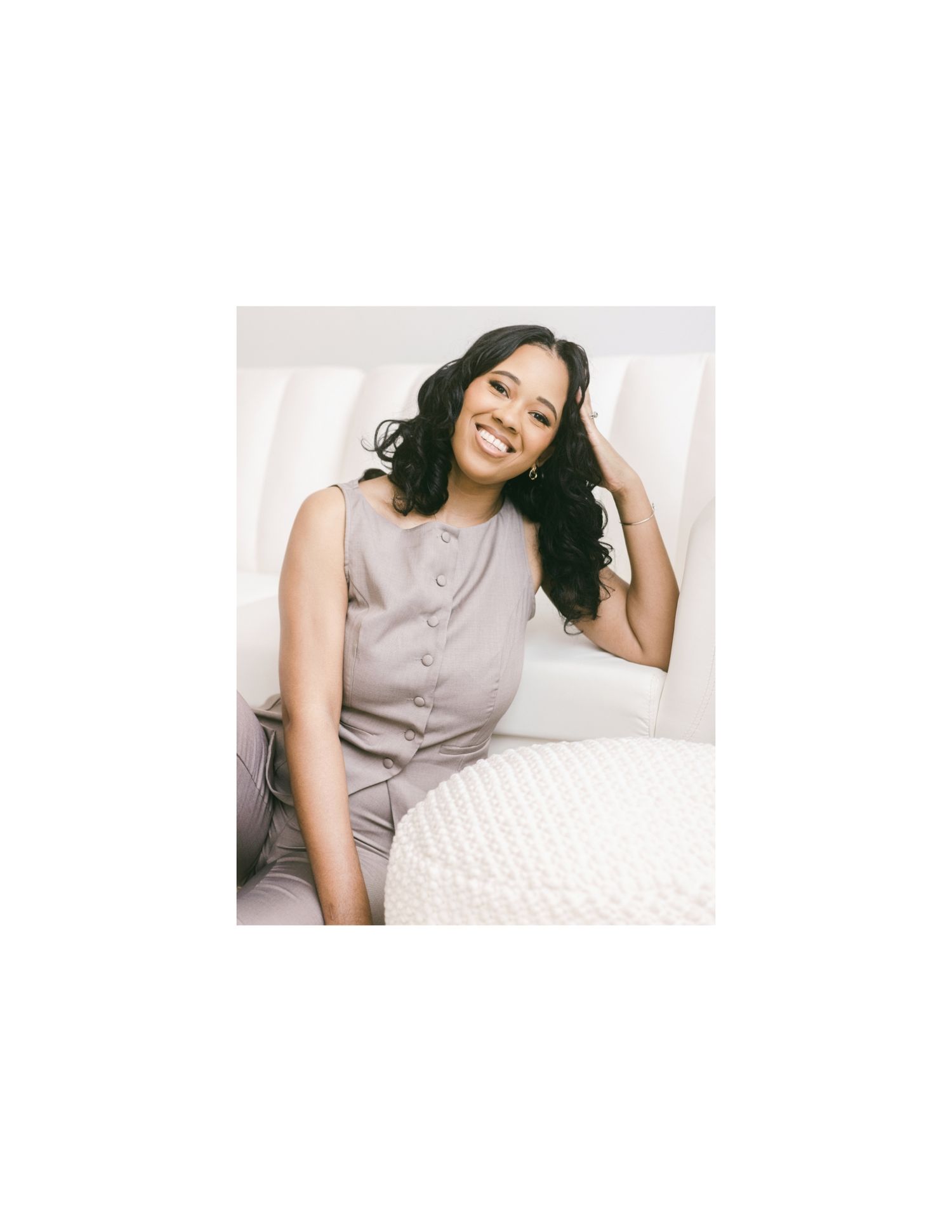 Smiling woman in gray button-up sleeveless top relaxing on a white couch, looking joyful.