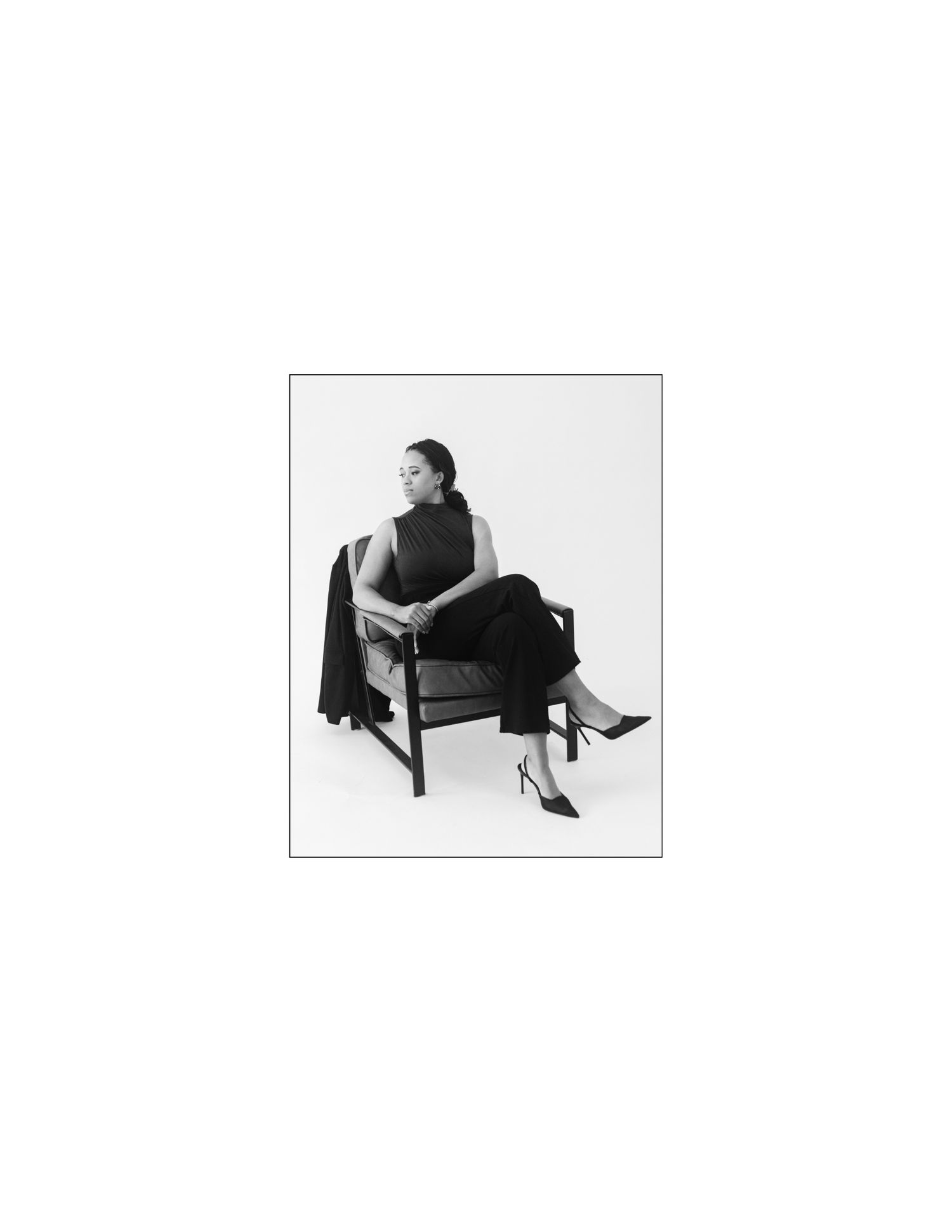 Woman in black outfit seated elegantly in a wooden chair, black and white fashion portrait photography.
