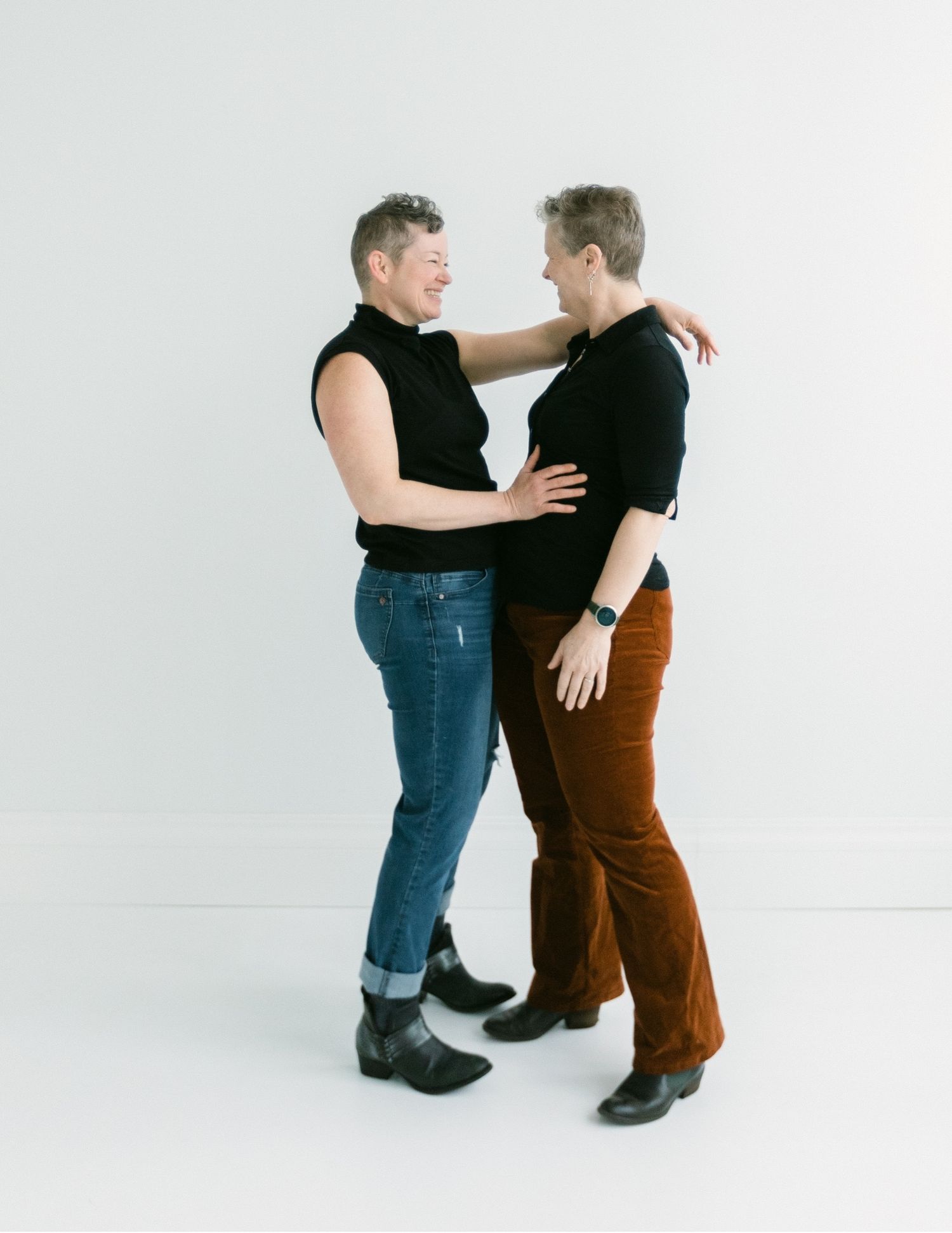 Two women in black tops and pants share a warm embrace, smiling at each other against a white background.