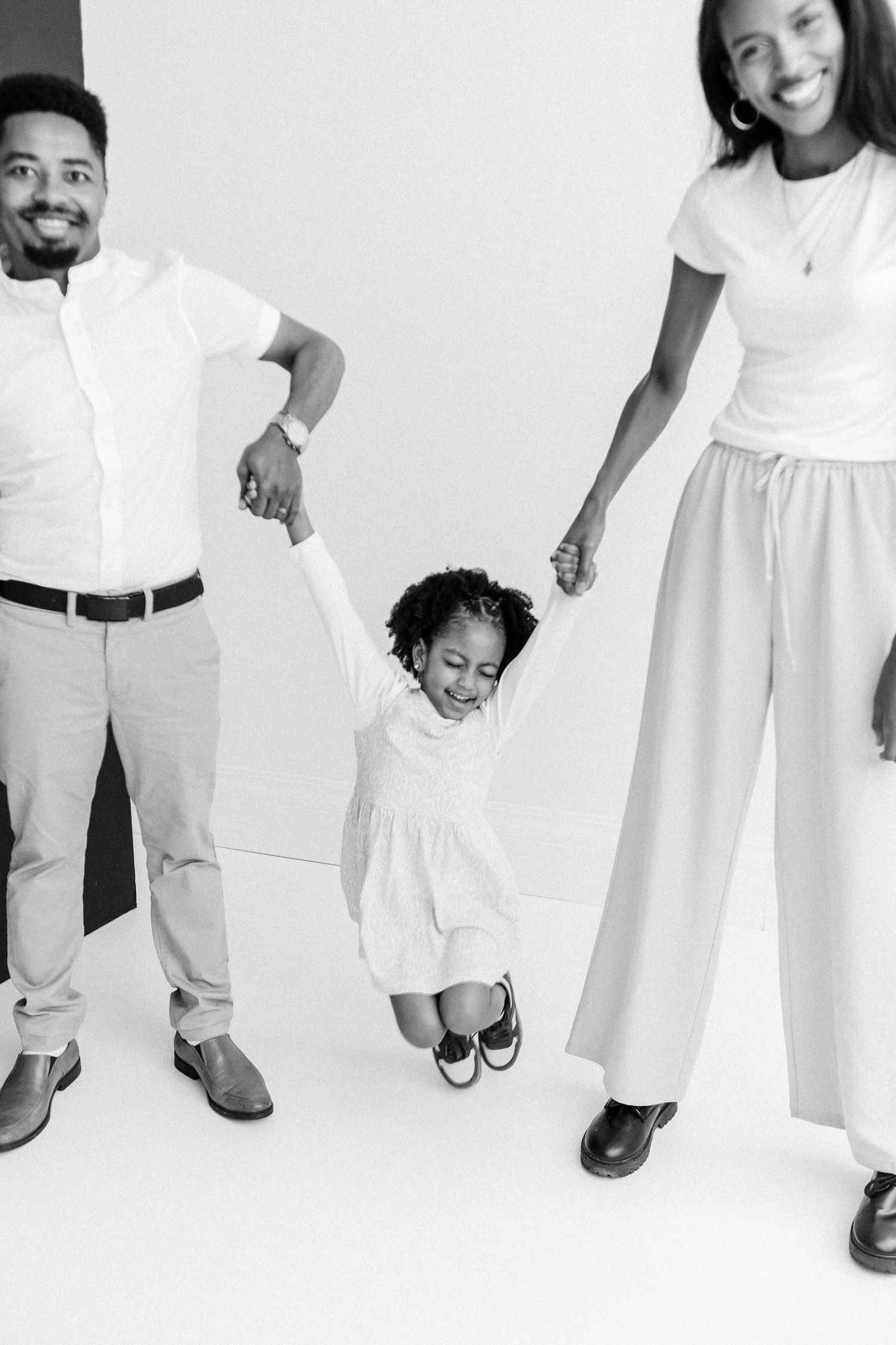 Black and white photo of parents lifting young daughter by hands in bright studio.