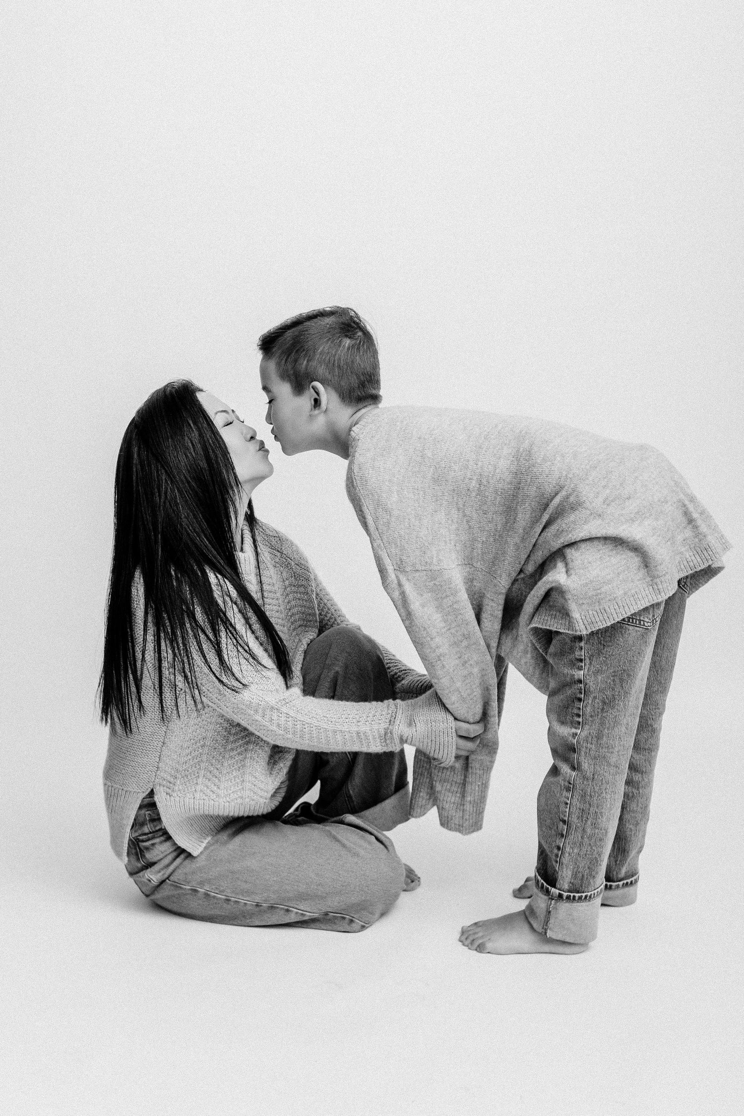 Black and white image of a mother sitting on floor receiving a kiss from her son leaning over her.