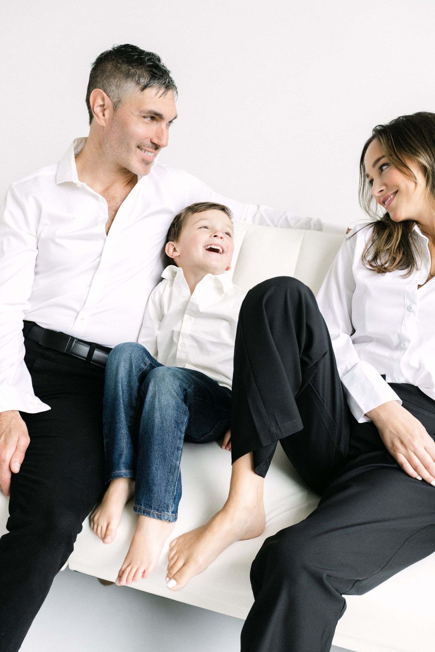 Happy family of three sitting on a white sofa, parents in white shirts smiling at laughing barefoot child.