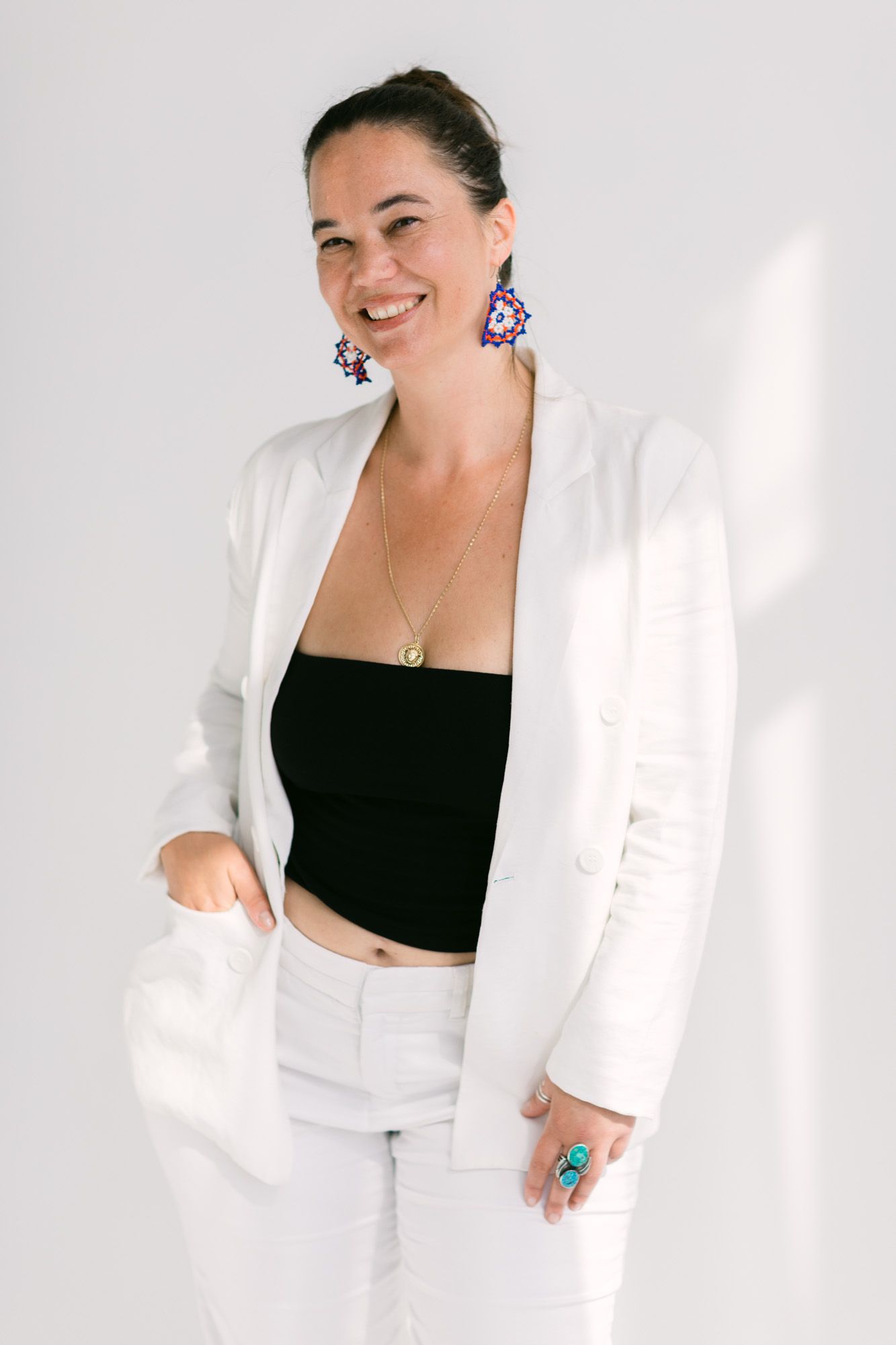 Smiling woman in white blazer, black crop top, and white pants, wearing colorful earrings and turquoise ring.