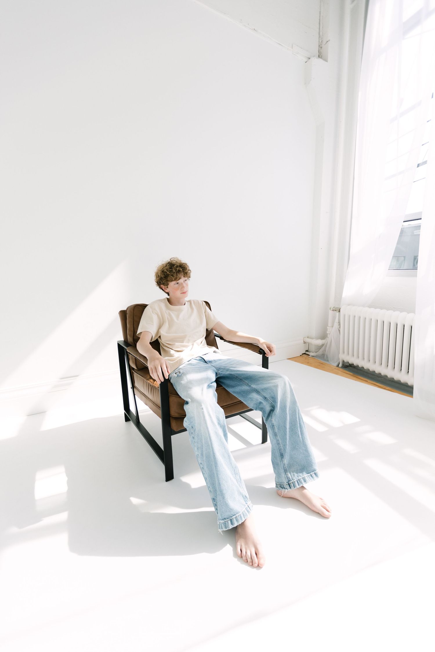 Young man with curly hair in beige tee and jeans sits barefoot in sunlit white studio chair.