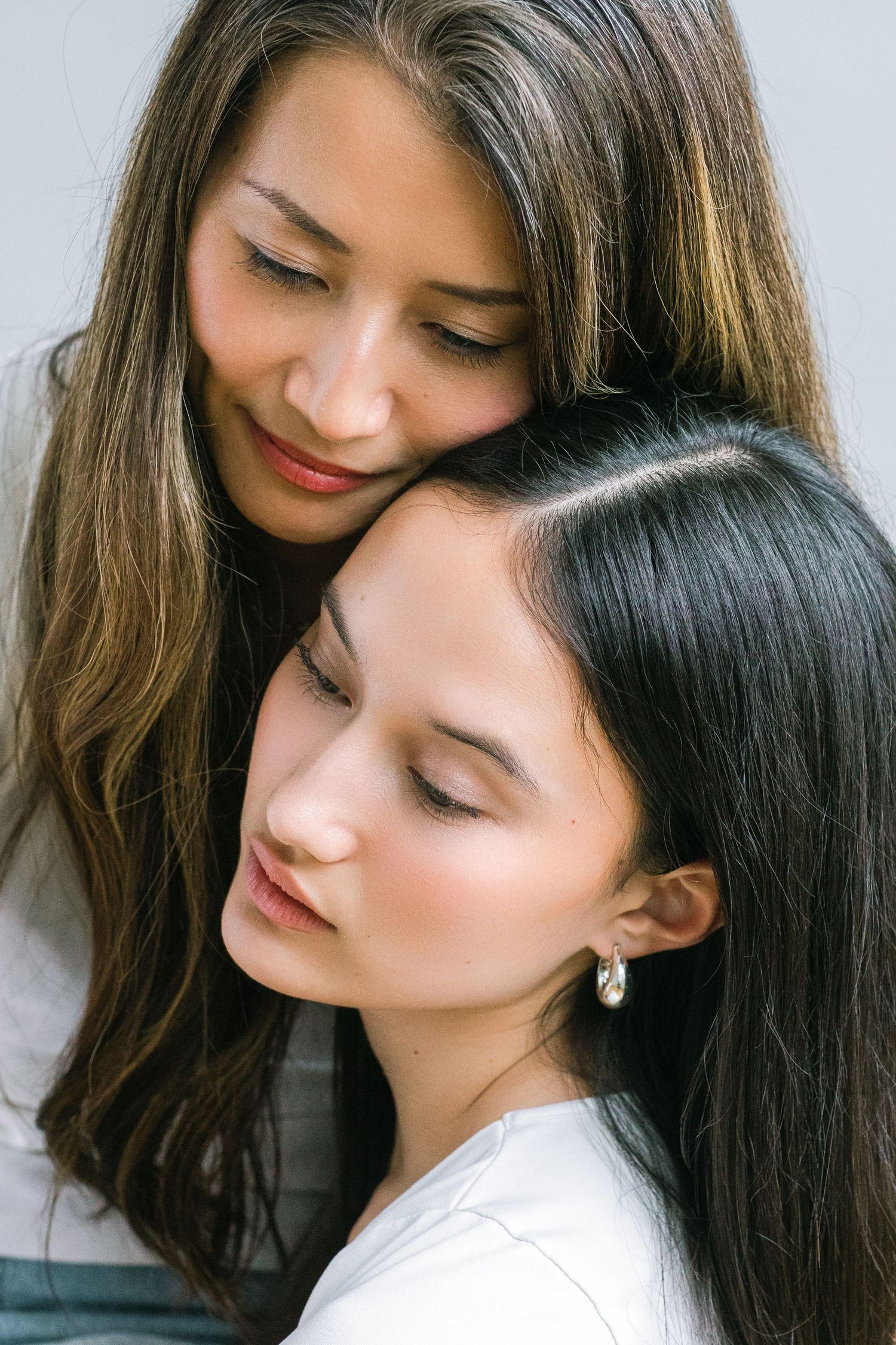 Two young women with long dark hair share a tender embrace, eyes closed, in a serene portrait photo.