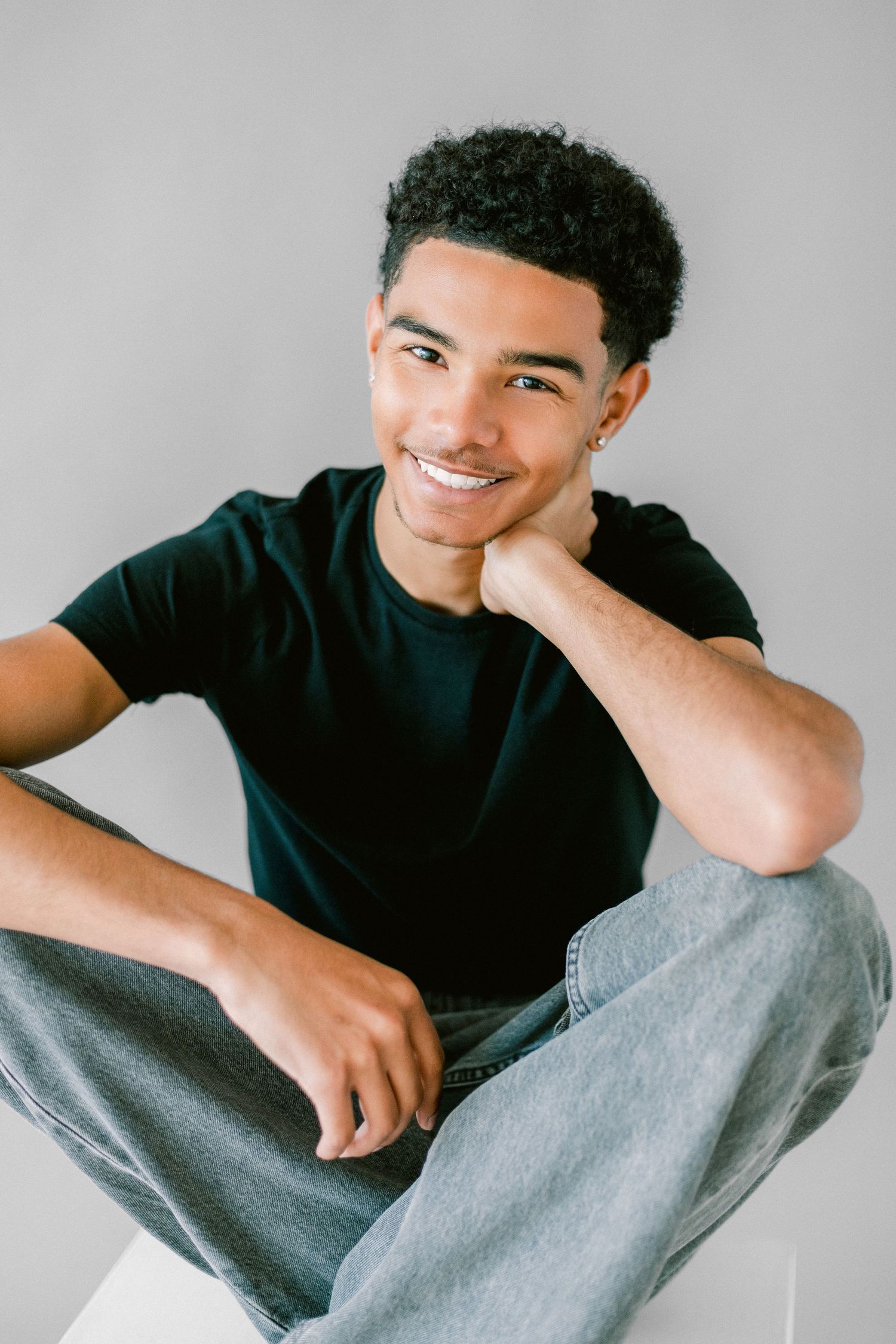Young man in black t-shirt and jeans sitting cross-legged, smiling with hand on chin against gray background.