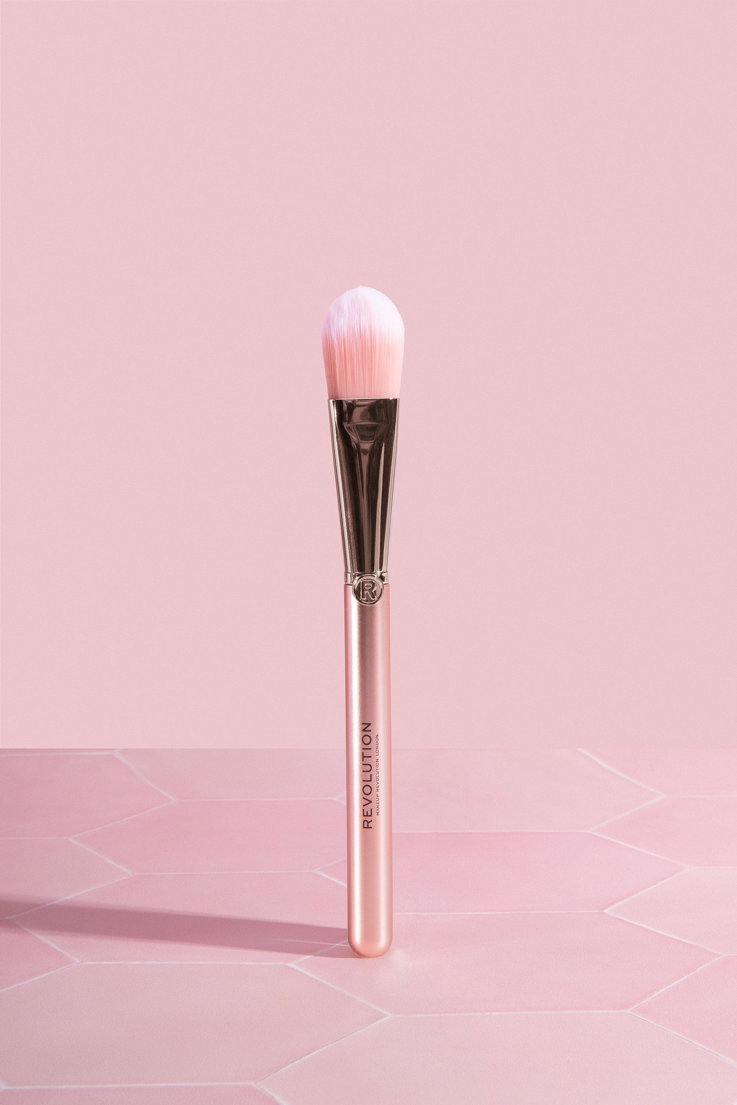 Revolution pink and rose gold makeup foundation brush standing upright against a soft pink geometric background.