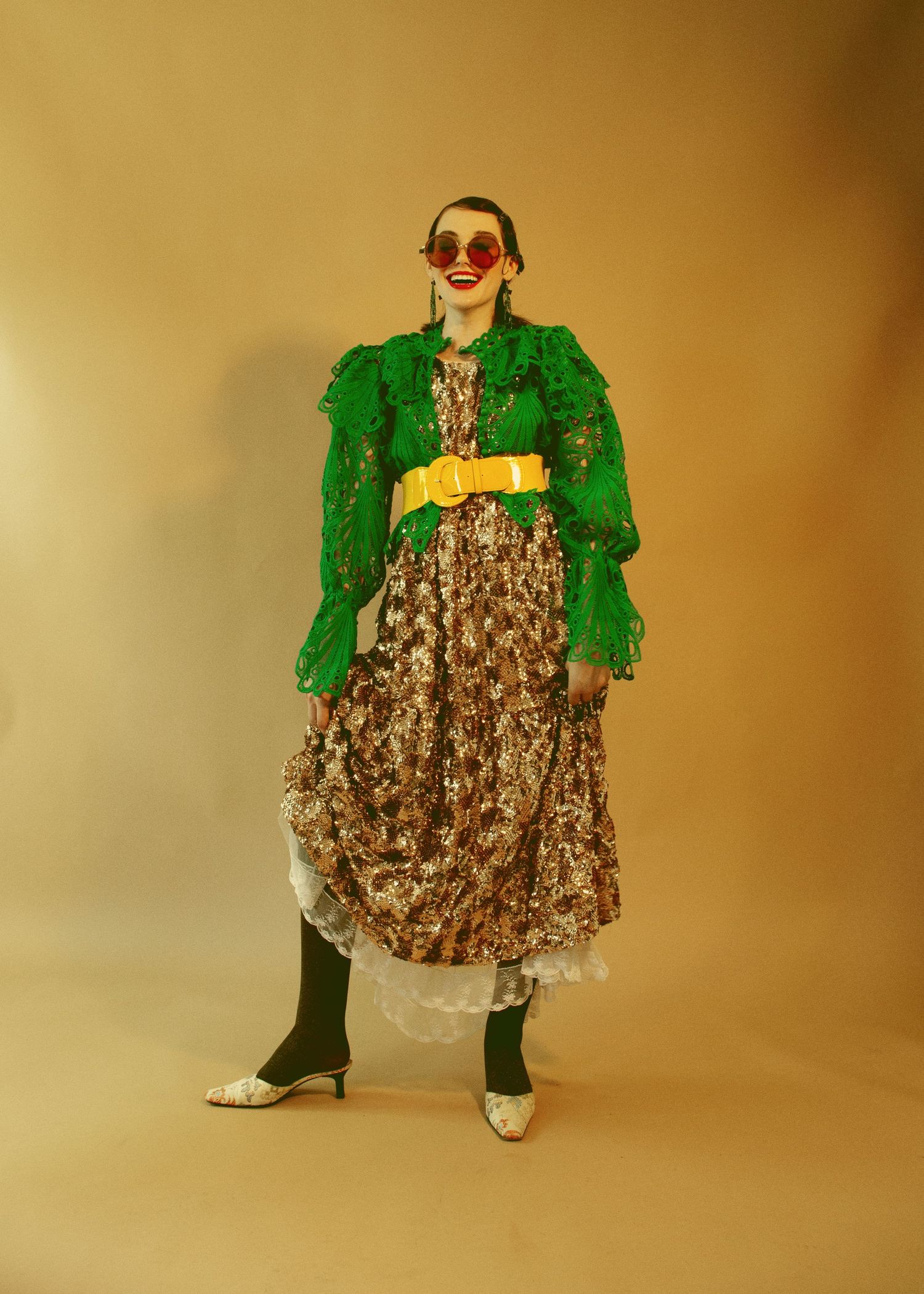 Model wearing green lace jacket, floral maxi dress, yellow belt, red sunglasses, and silver heels in studio.