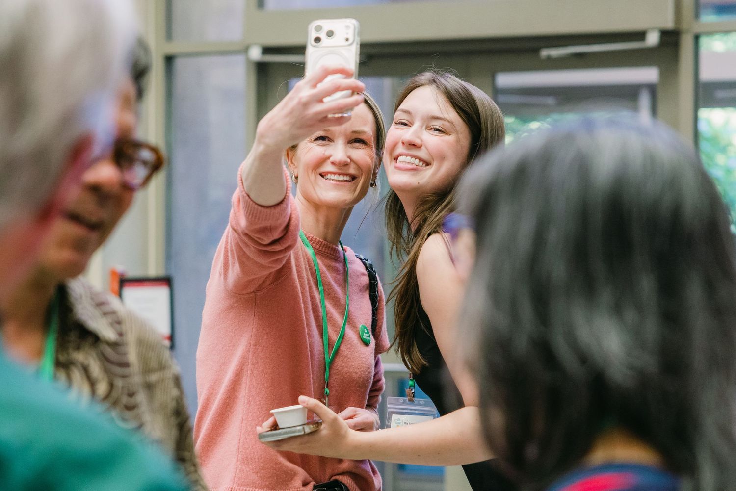 Two women taking a selfie with smartphone at a professional event while wearing lanyards.