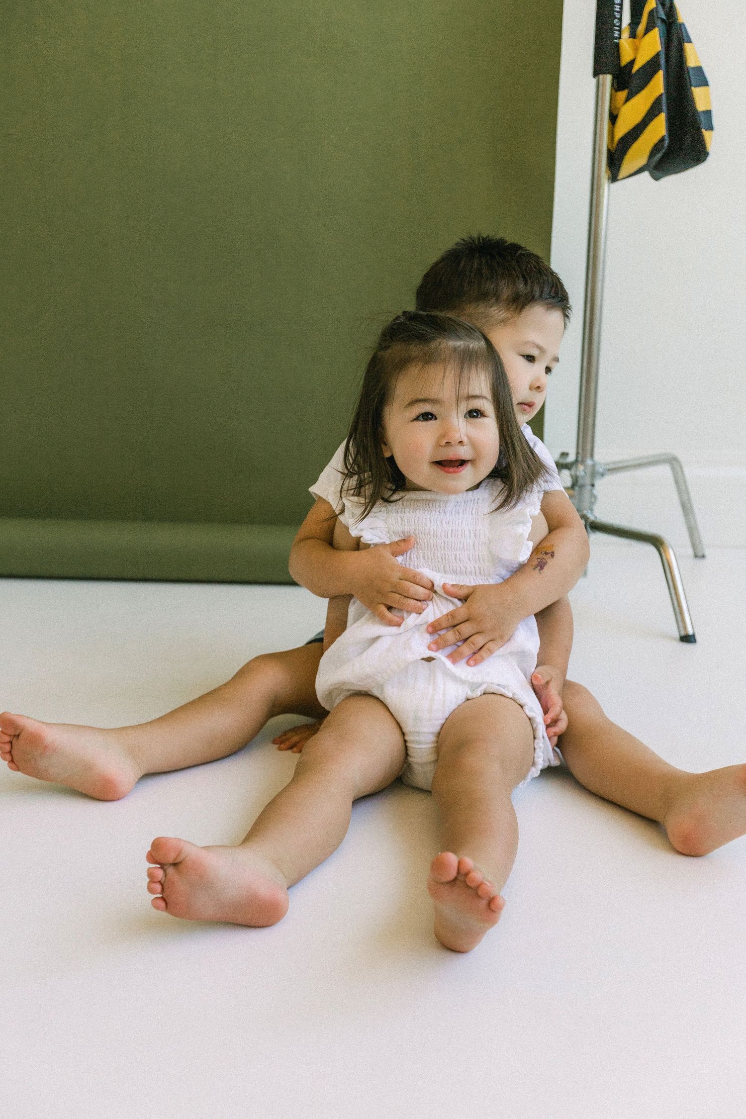 Two young Asian children sitting on white floor, older boy hugging smiling baby girl in white outfit.