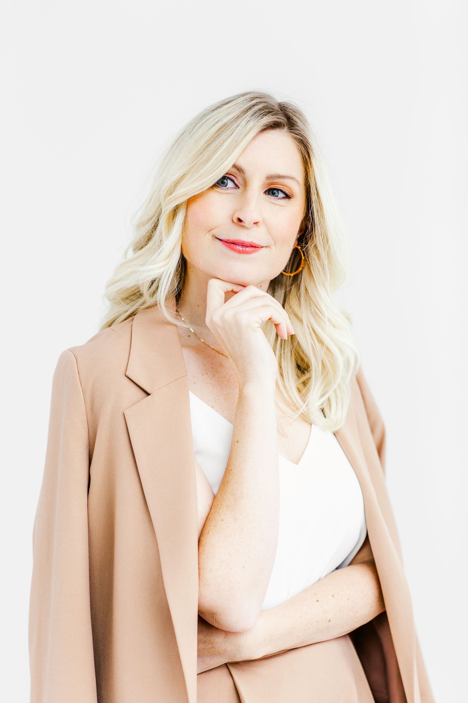 Confident blonde woman in beige blazer and white top posing thoughtfully against a light background.