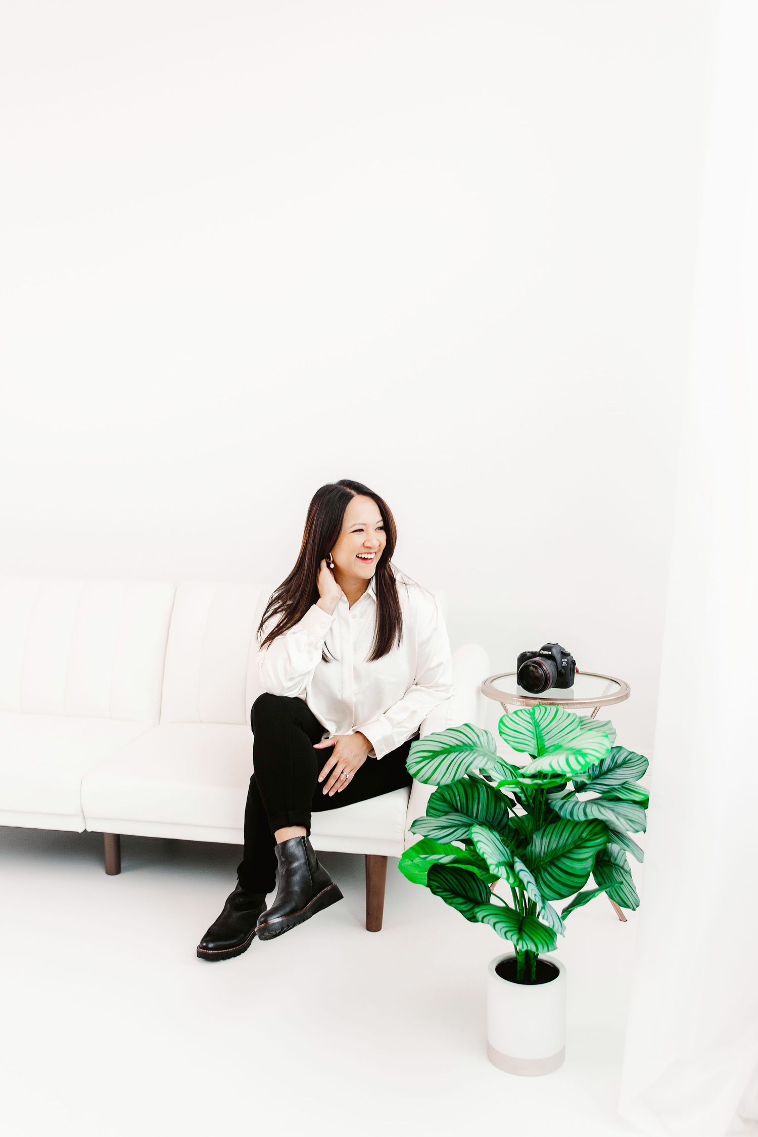 Woman in white shirt and black pants laughing on a white sofa beside a green plant and camera.