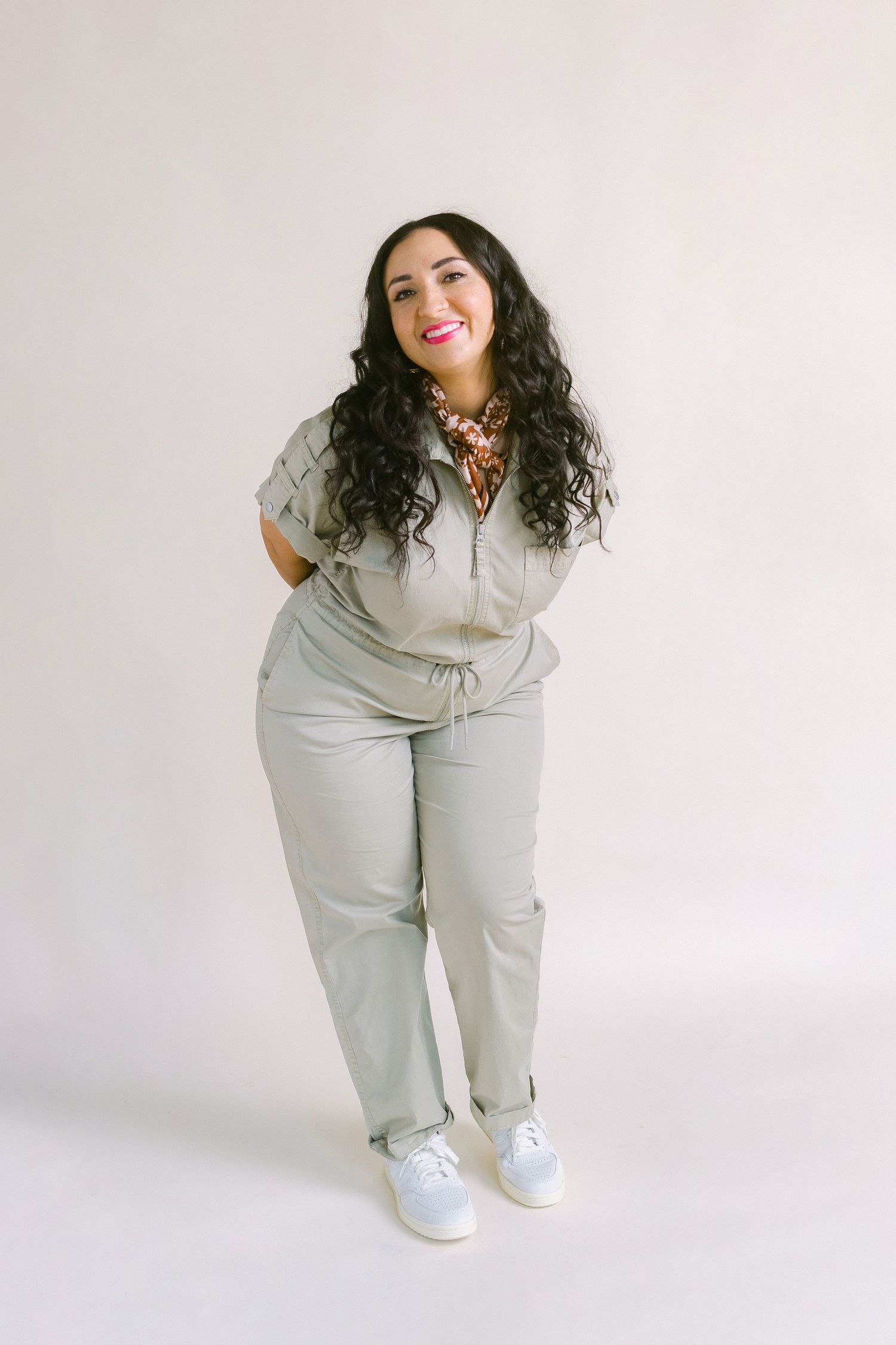 Woman in sage green jumpsuit with white sneakers and floral necklace, smiling against white background.