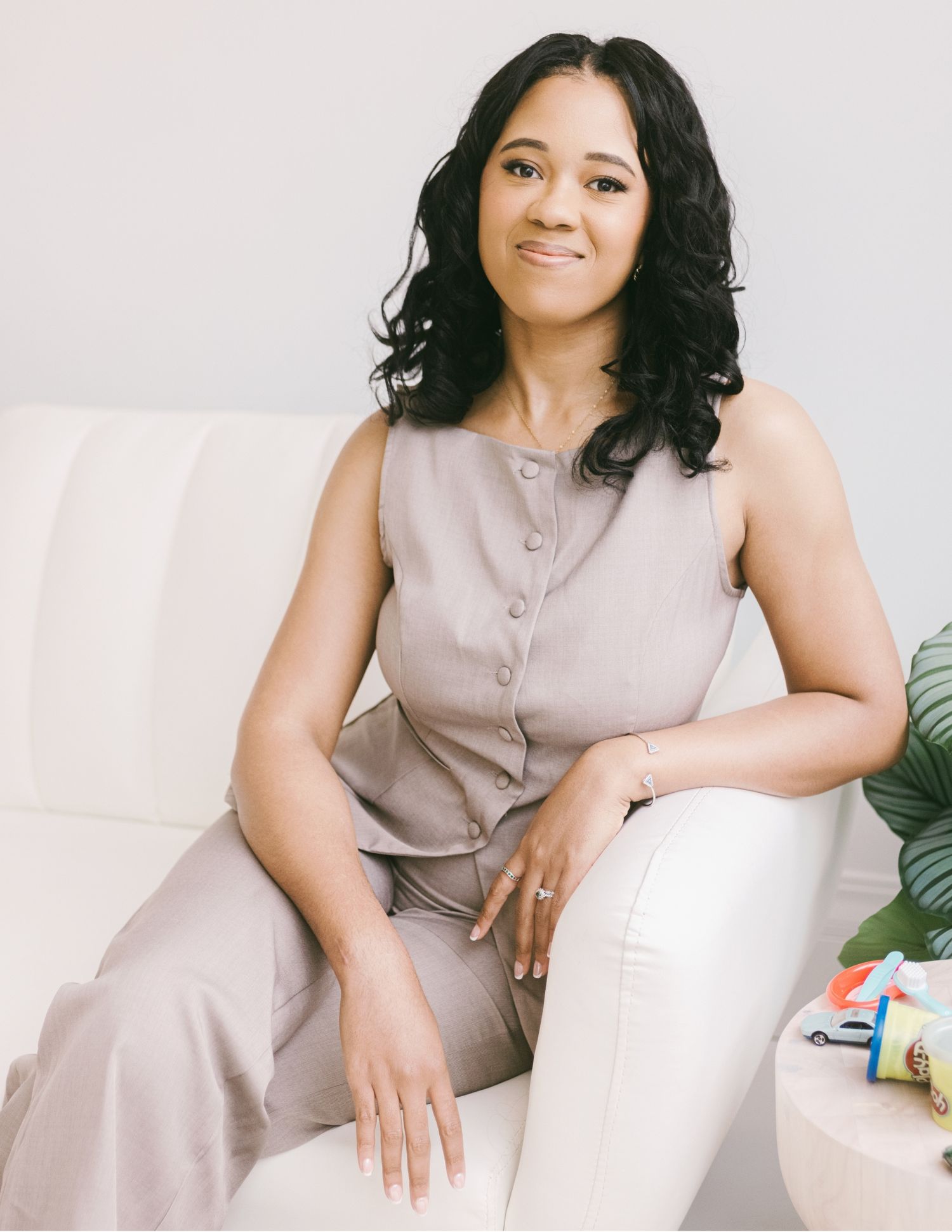 Woman in gray sleeveless top and white pants sitting gracefully on a white sofa, smiling warmly.