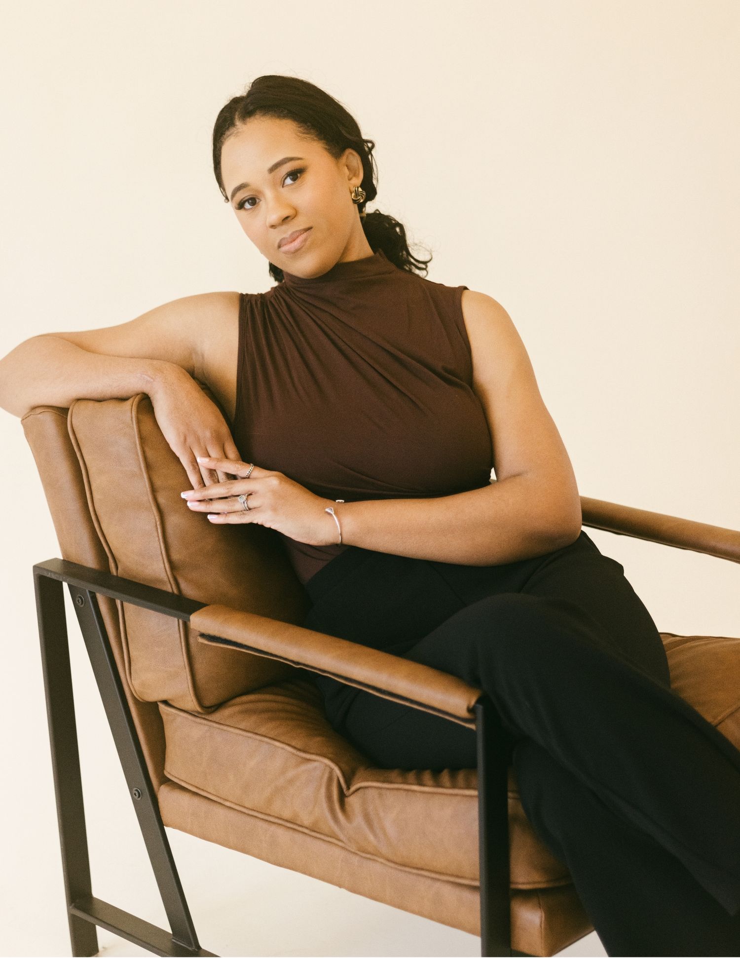 A woman in a brown sleeveless top and black pants sits relaxed in a tan leather chair against a neutral background.