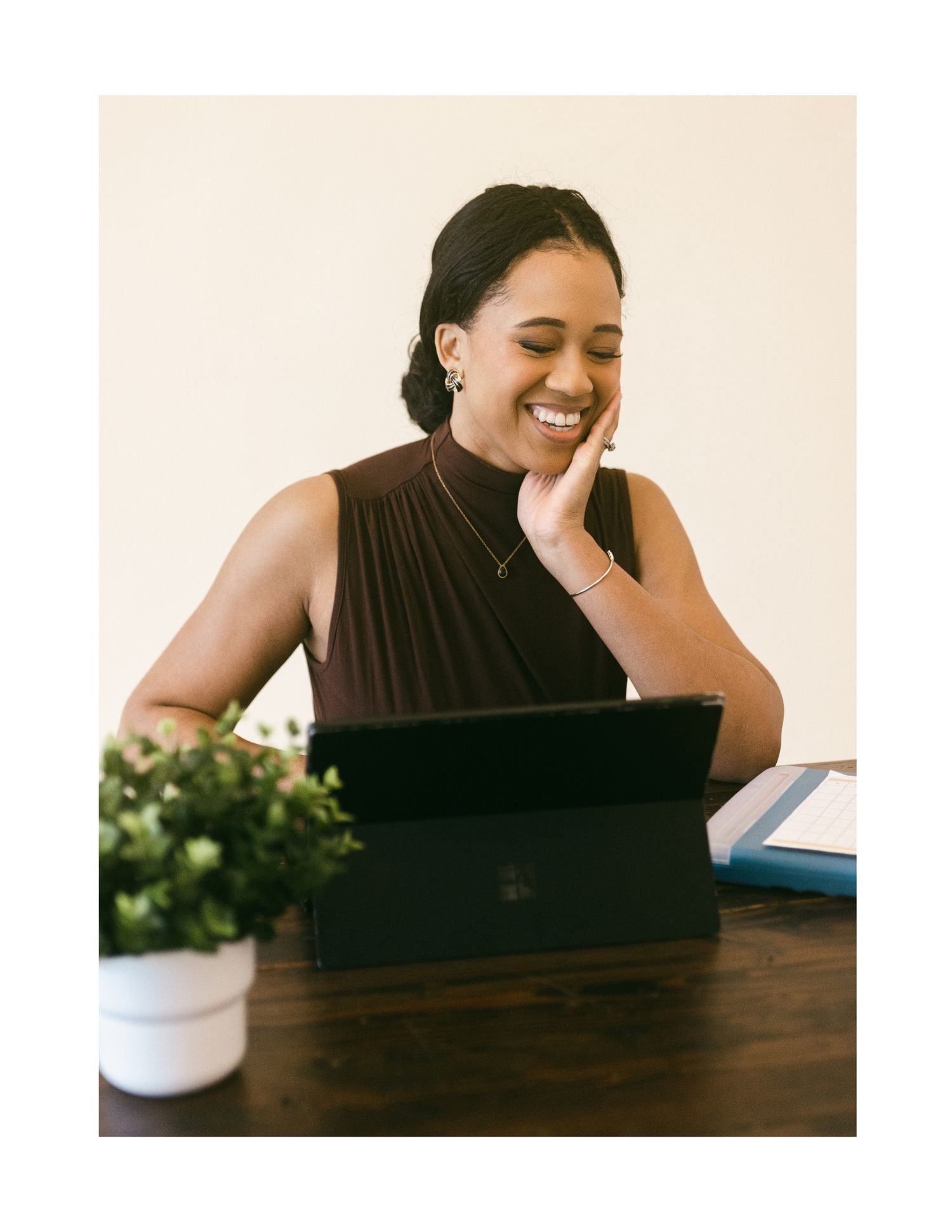 Smiling young woman in brown sleeveless top sitting at wooden desk with tablet, plant nearby, bright background.