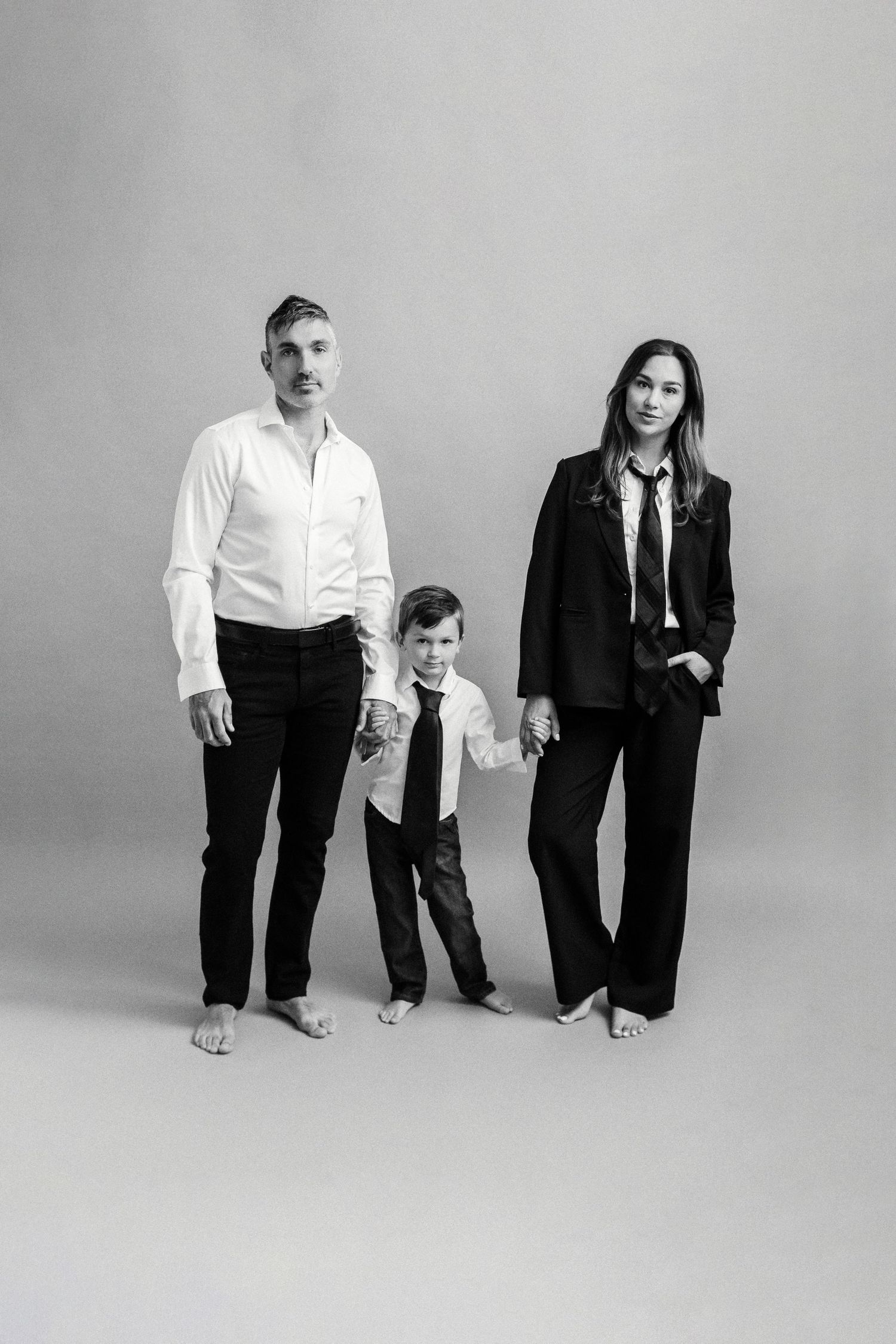 Black and white family portrait of two adults and a child in formal attire, holding hands against a neutral background.