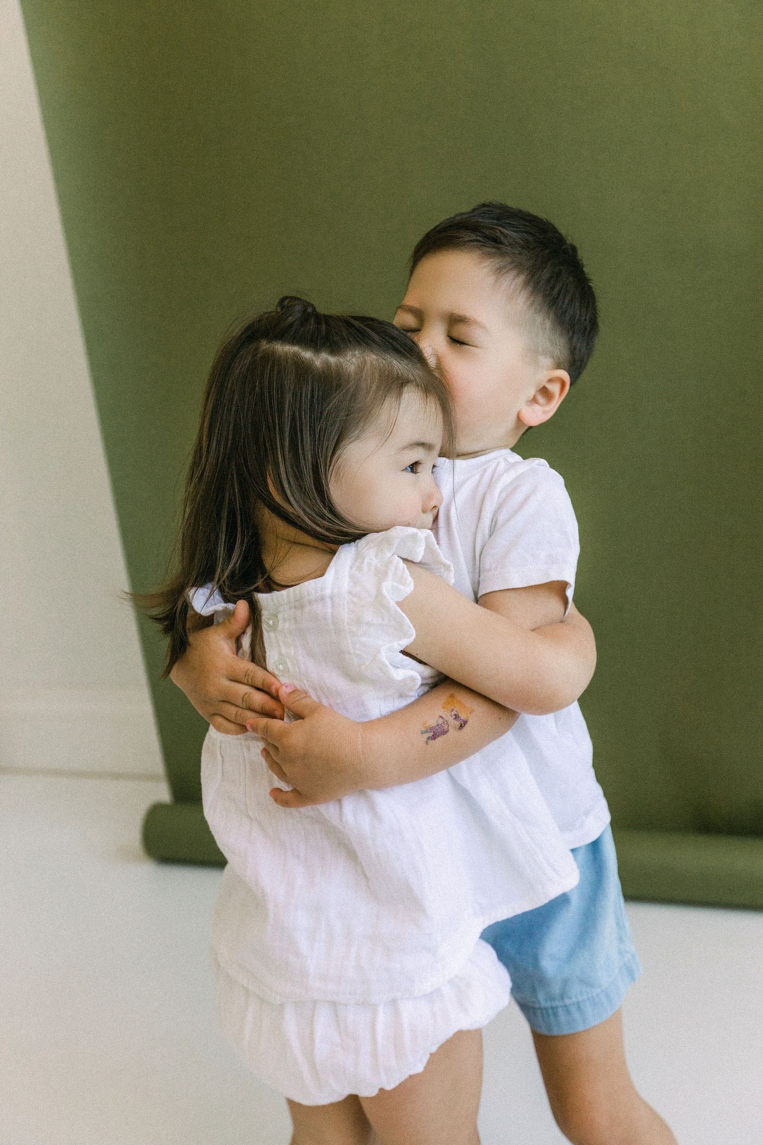 Two young Asian siblings share a sweet hug and kiss, dressed in white and blue outfits against a green backdrop.