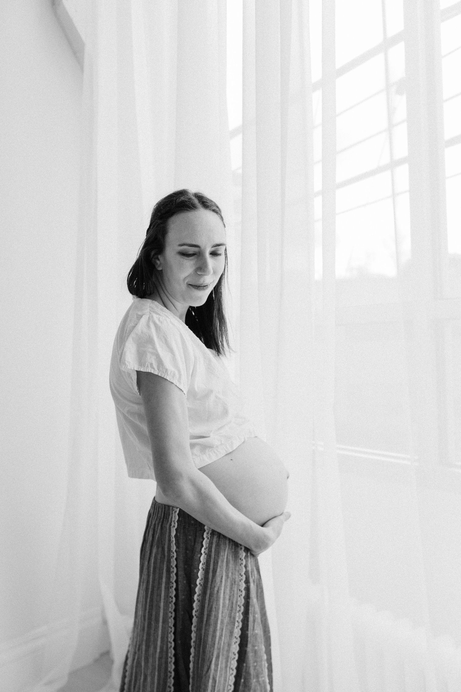 Black and white photo of a pregnant woman in white top and skirt, cradling her baby bump by a bright window.