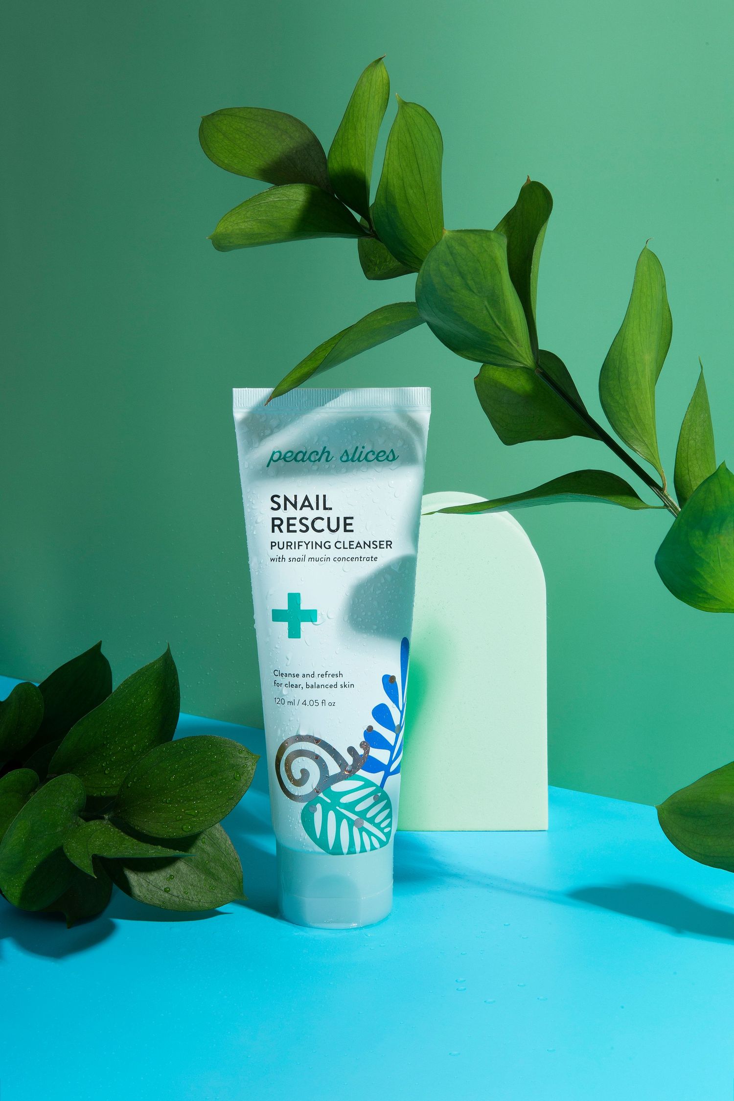 Peach Slices Snail Rescue Purifying Cleanser tube displayed with green tropical leaves on teal background.