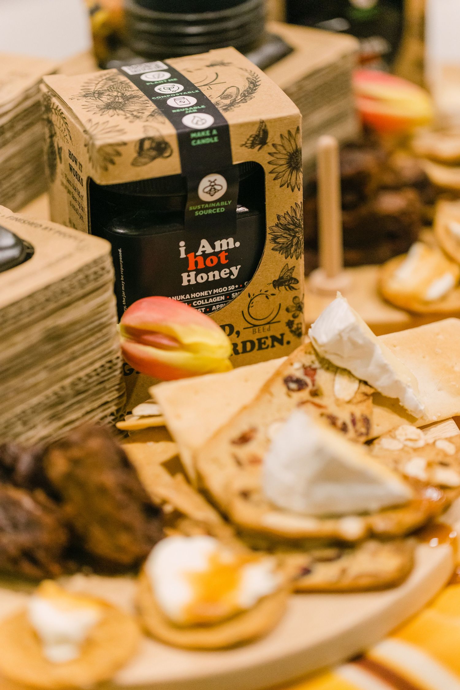 Charcuterie board with brie, crackers, and apple slices beside a jar of I Am Hot Honey and branded packaging.