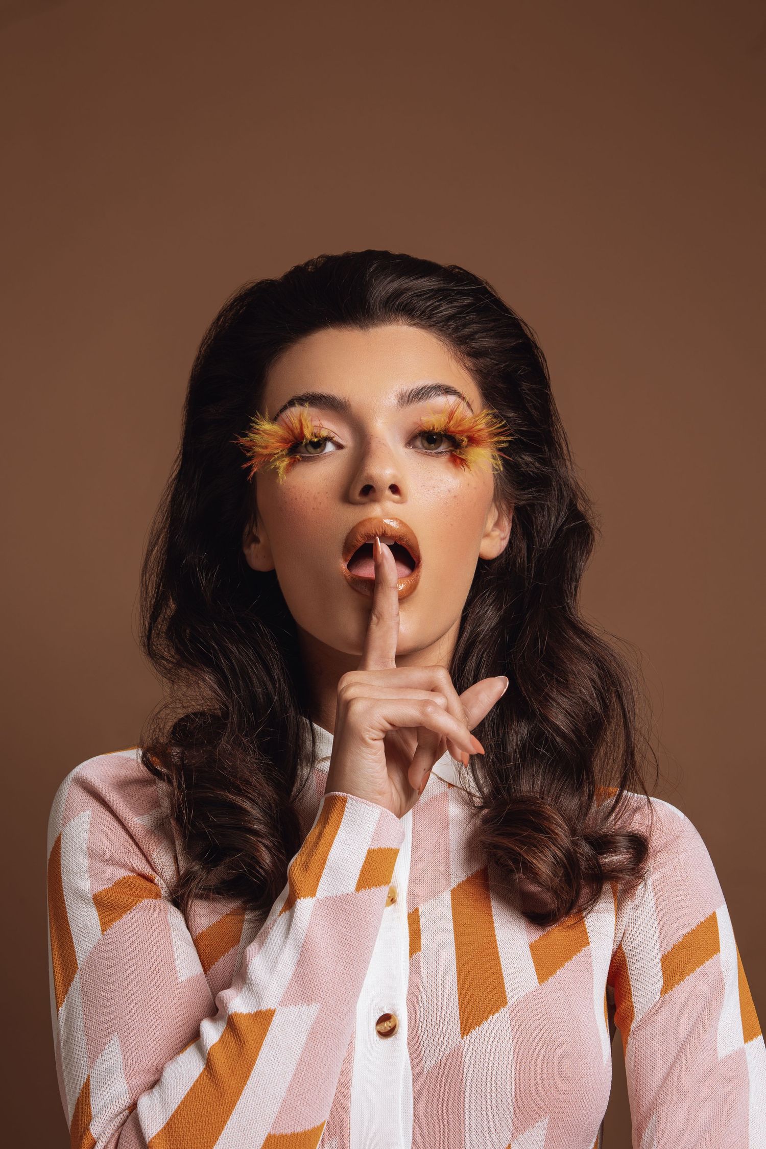 Brunette woman with feathered orange lashes in striped shirt holds finger to lips on brown background.