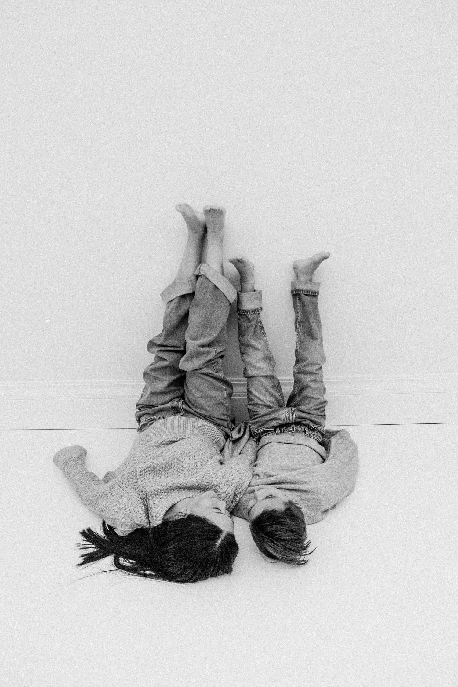 Black and white photo of two children lying on floor with legs up against a white wall.