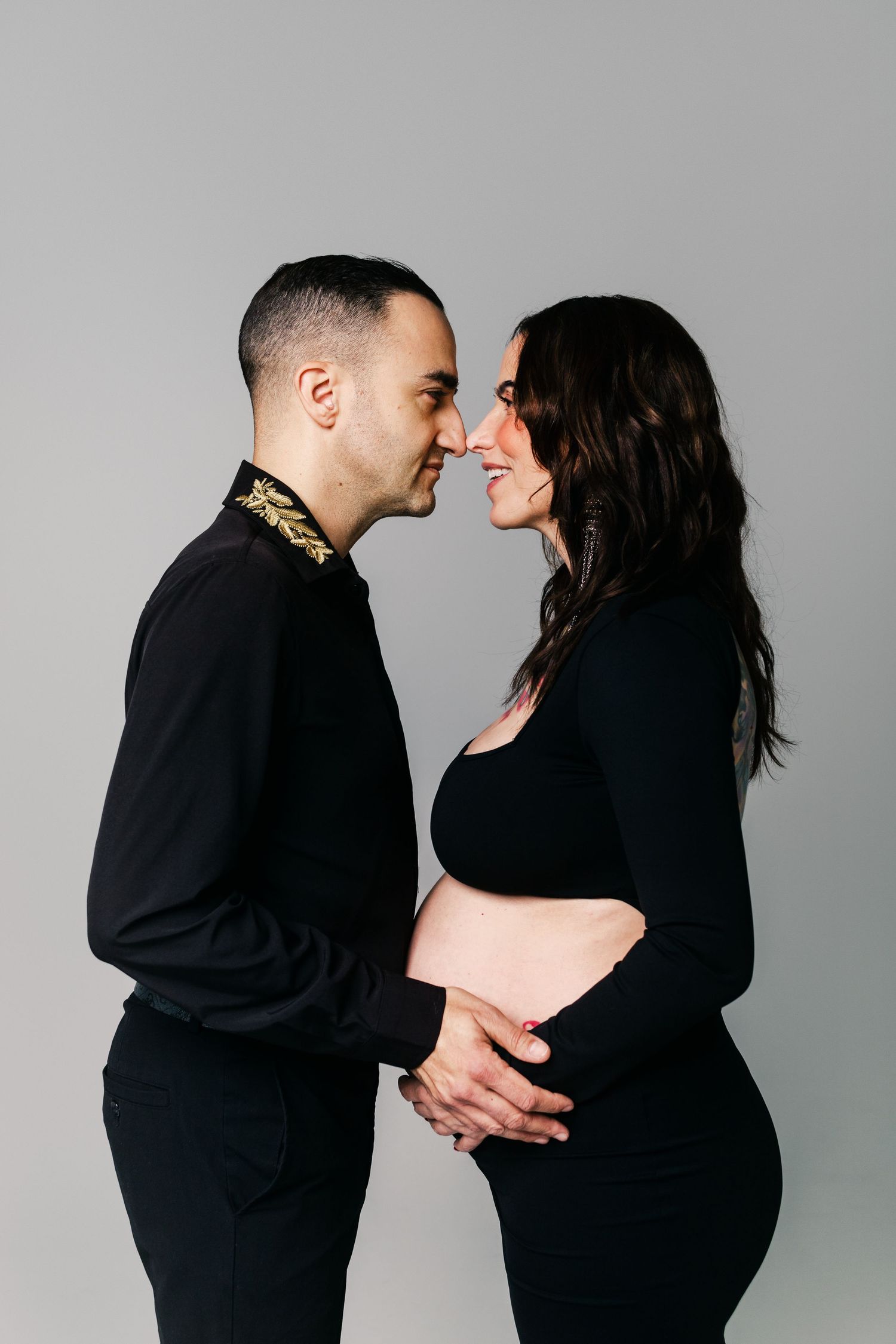 Couple in matching black outfits touching foreheads tenderly, hands cradling pregnant belly, gray background.