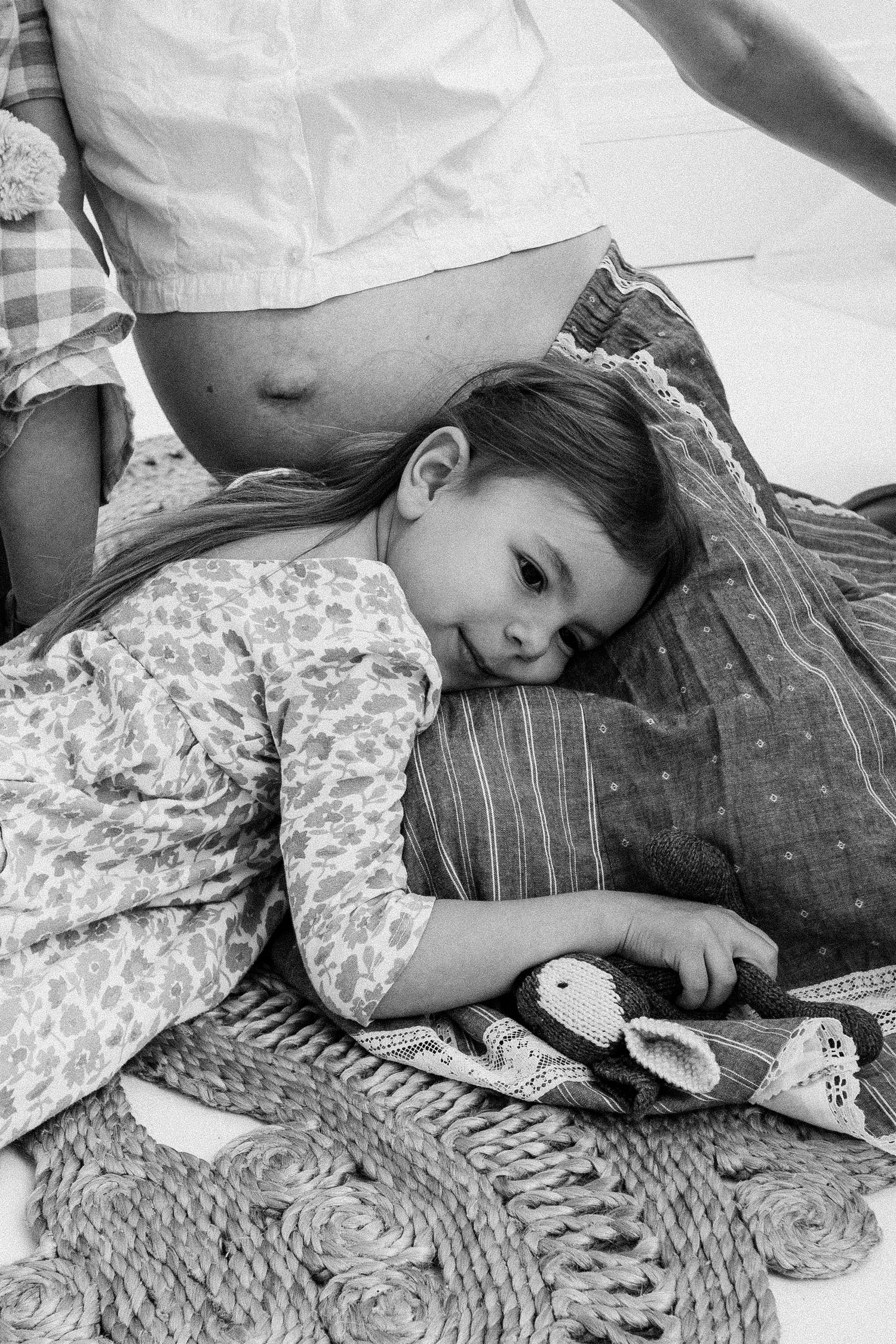 A young girl resting her head on pillows near a pregnant woman's belly in a black and white photo.