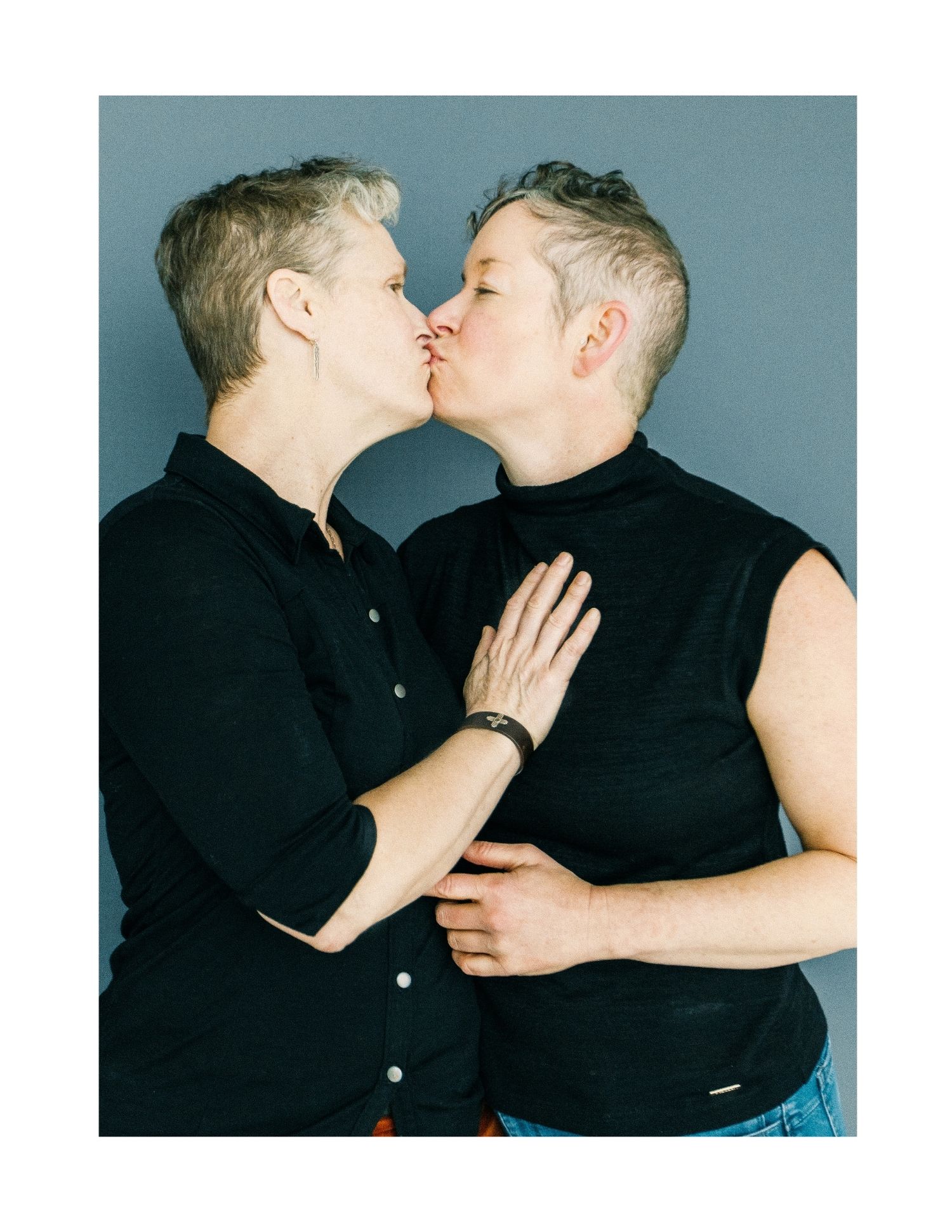 Two short-haired women sharing a tender kiss, embracing each other against a gray backdrop.