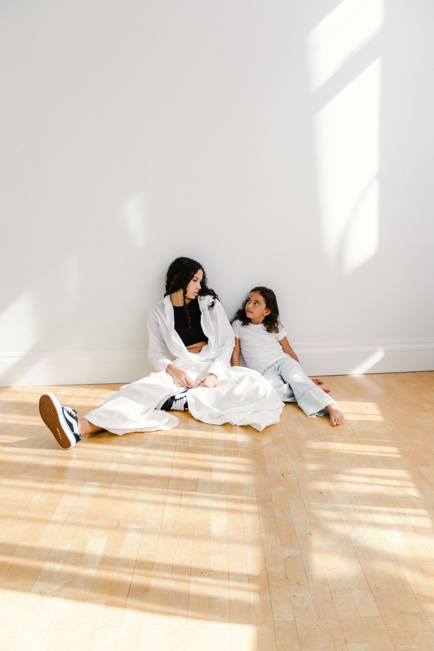 Woman and young girl sitting on hardwood floor against white wall in bright natural light, wearing casual white outfits.